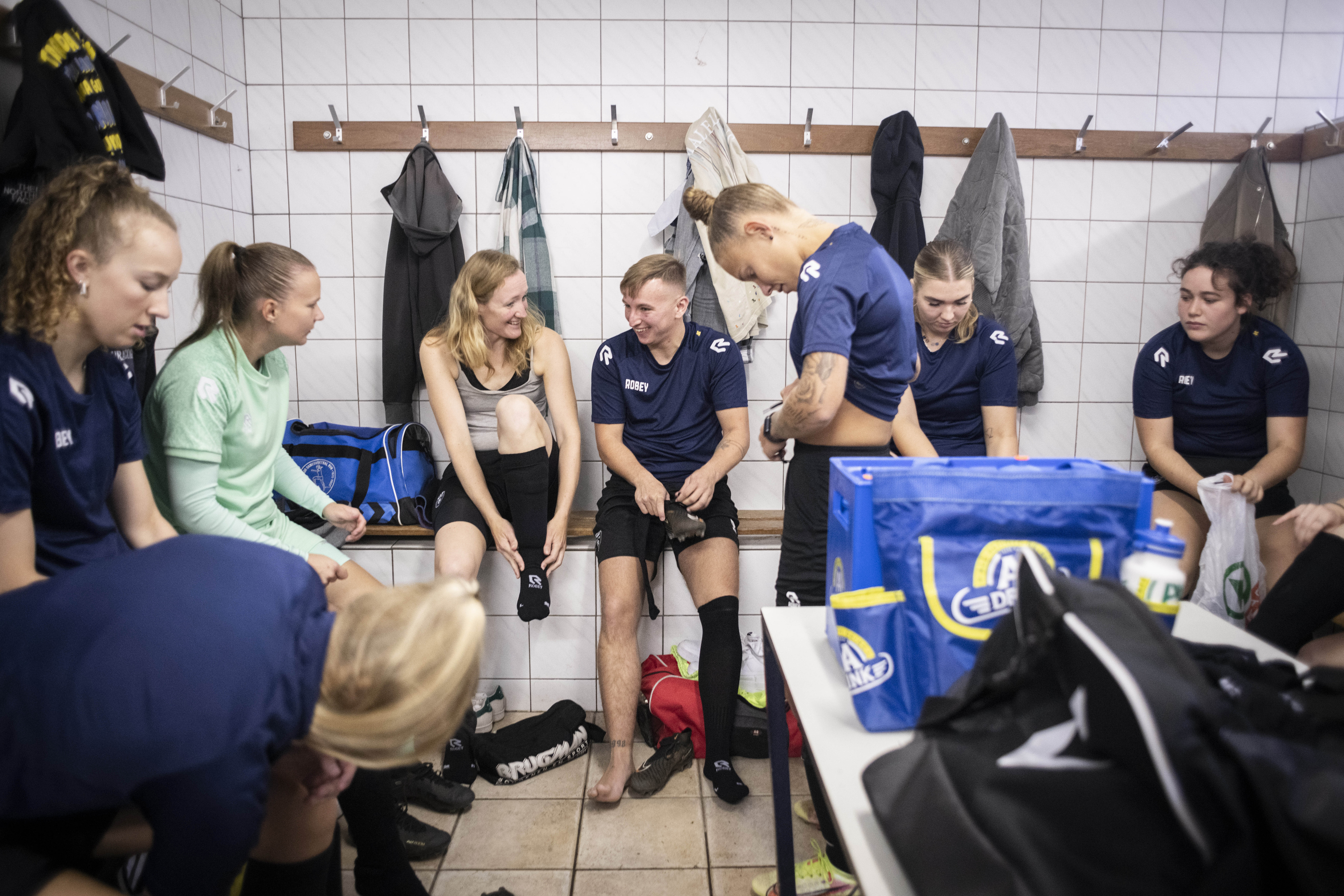 a football team changing in a locker room, putting their dark blue and black jersey while chatting. There are clothes and sports bags everywhere