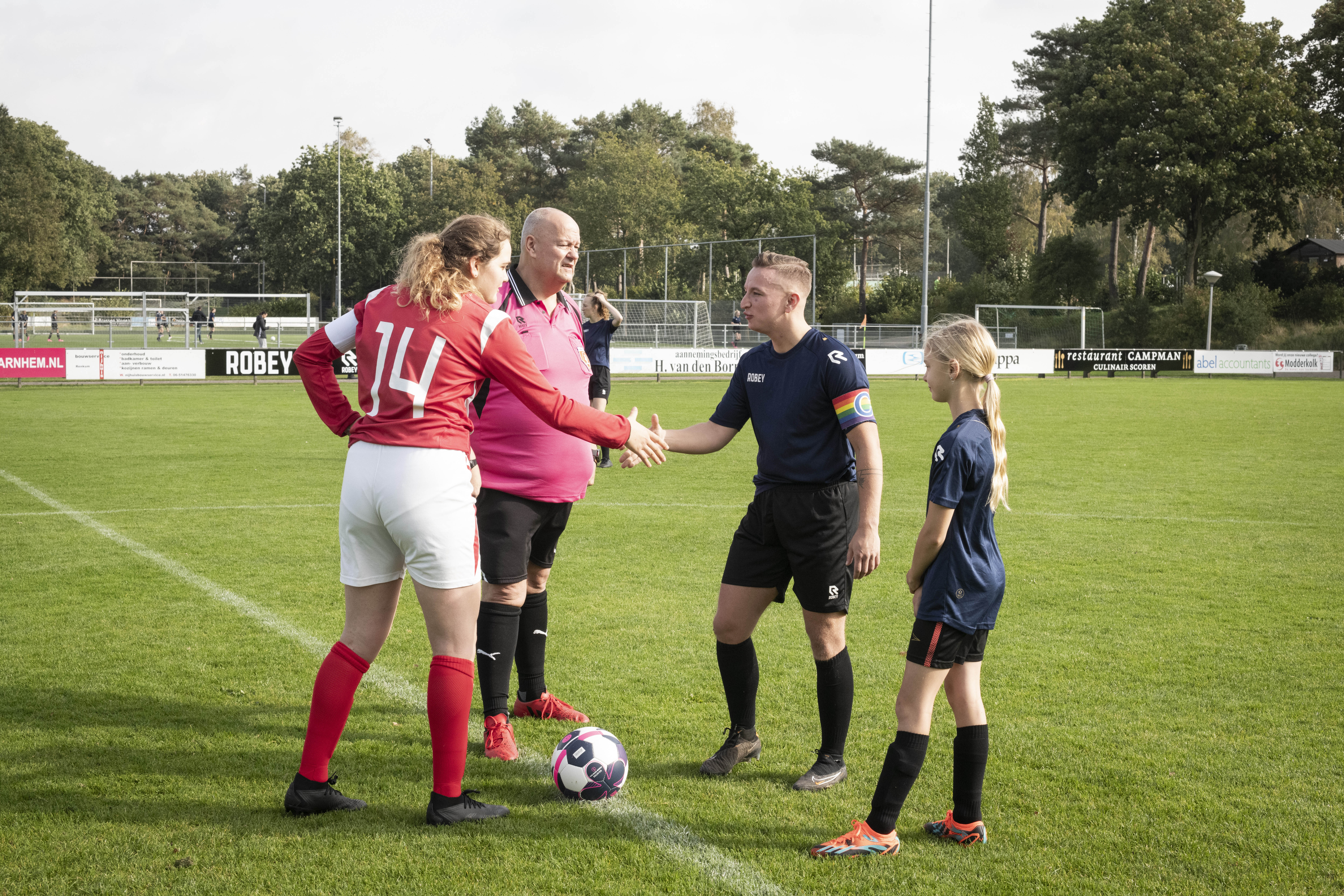 two football players shaking hands on the pitch next to the referee and a younger player. Two of them wear a dark blue and black jersey, the opponent wears a red and while jersey, and the referee is in black and red