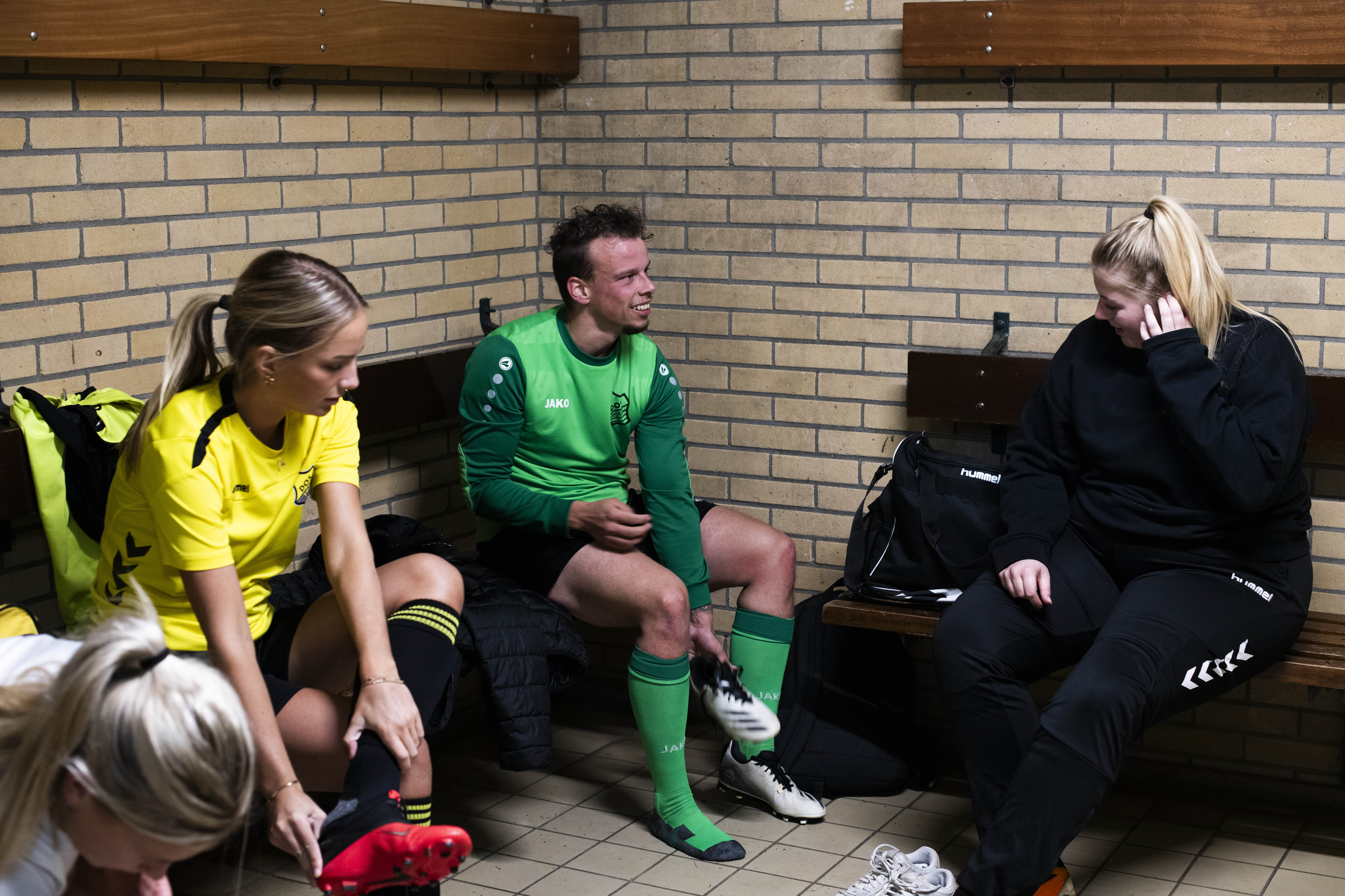 four players putting their sneakers on in a sports locker room. One of them is dressed in black, another in green, one in yellow, and the last one if white