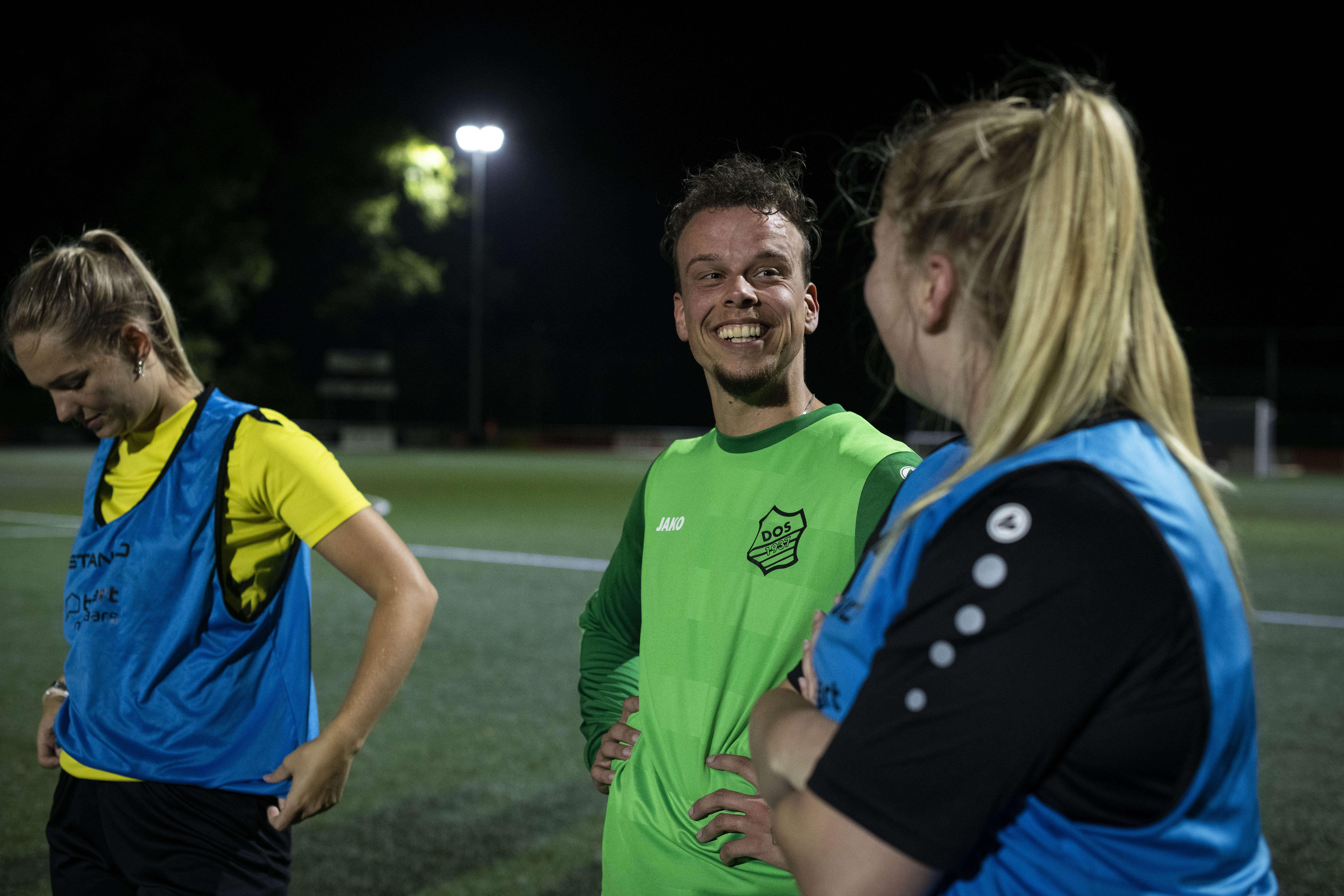 three players on a football field at night. Two of them look at each other smiling and the third one looks down. They're dressed in black, blue, yellow and green