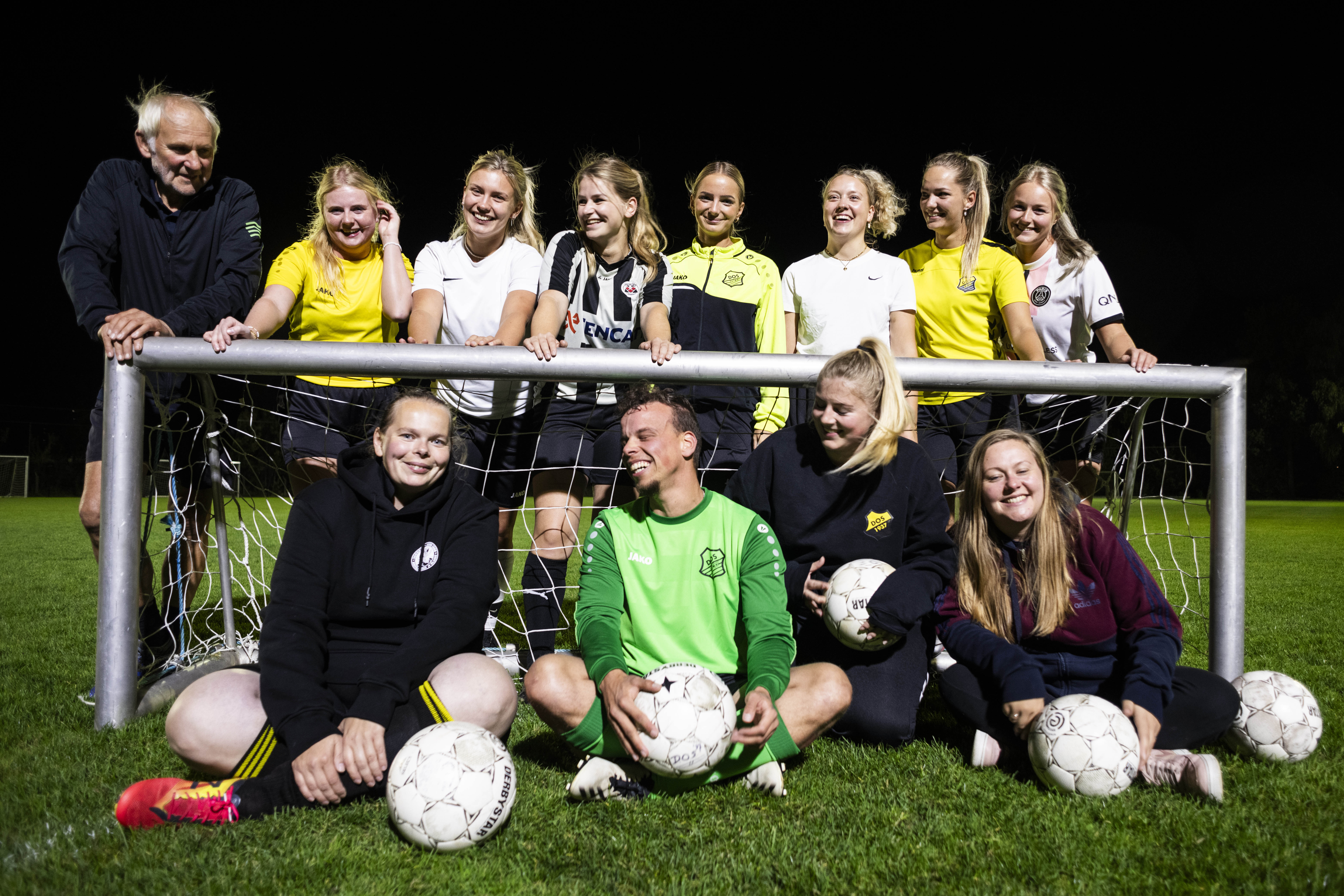 group photo of a women and queer football team near a small goal at night, they're wearing black, white and yellow football jerseys except for the goal keeper who's in green 