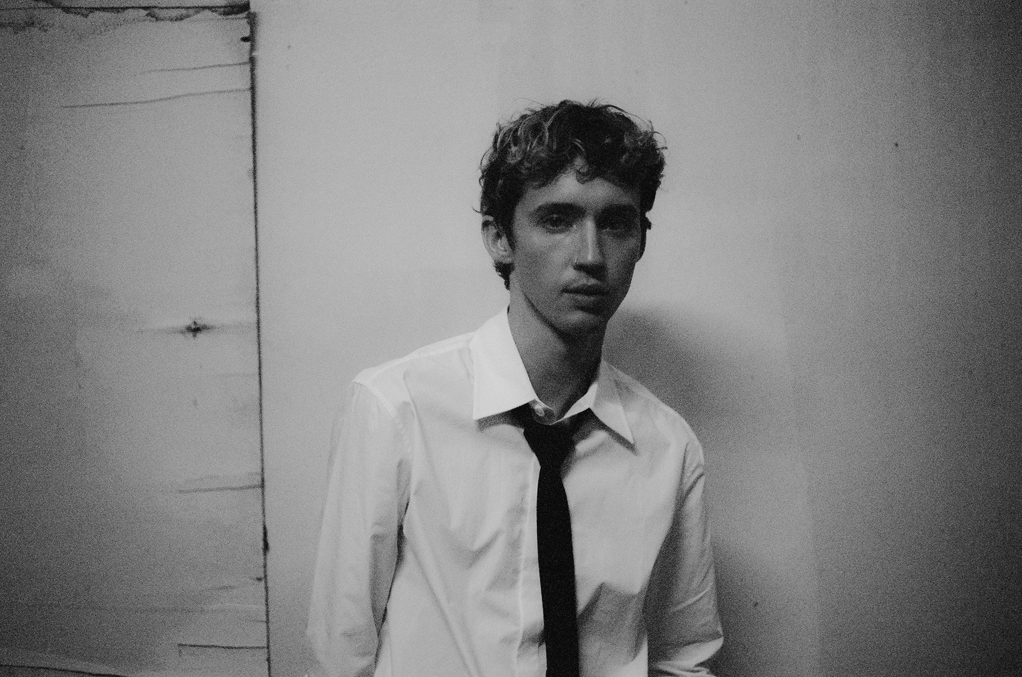 Troye Sivan stands against a wall wearing a shirt and loose-fitting tie