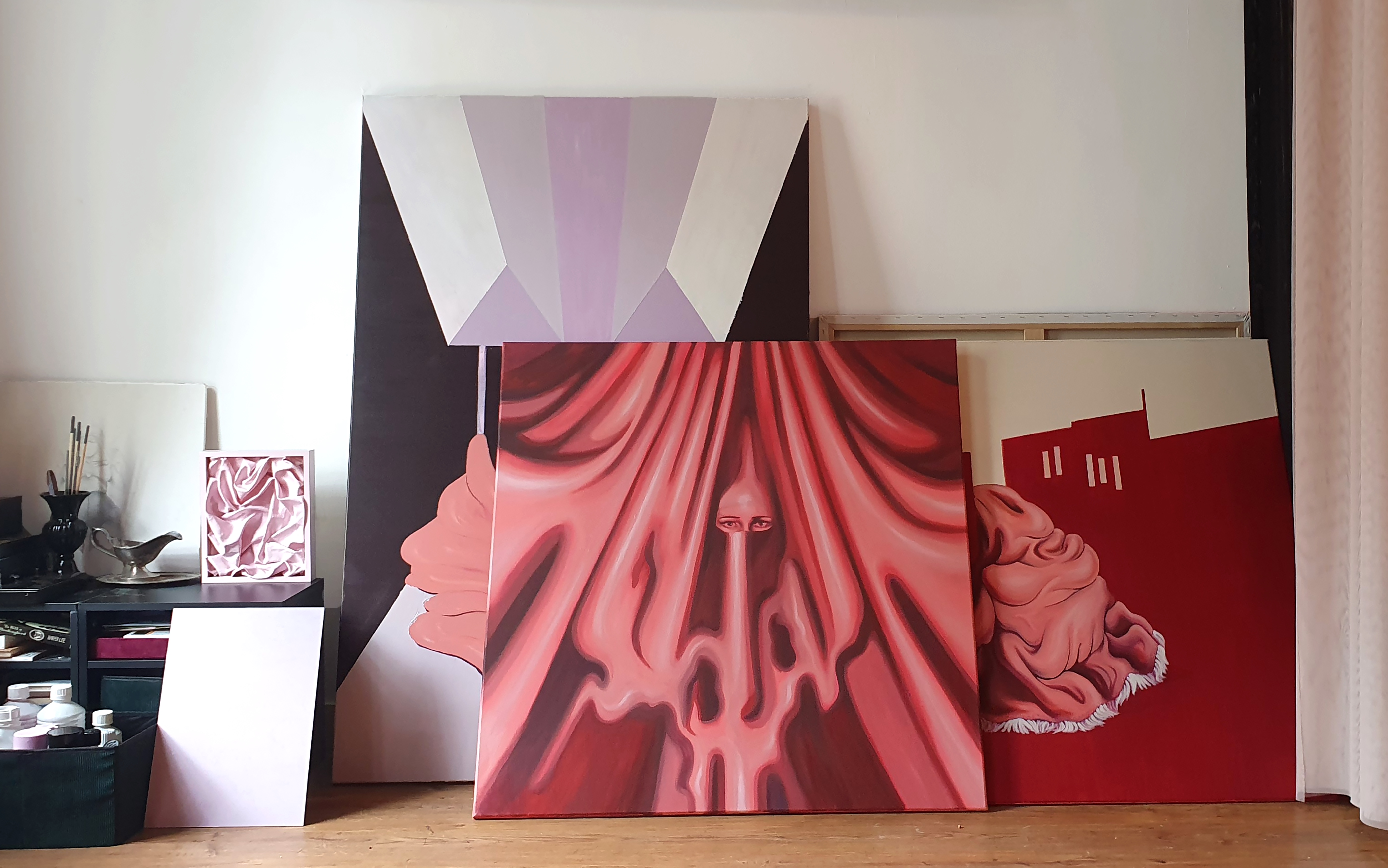 Photo of several pink, red, purple and black paintings against a white wall.