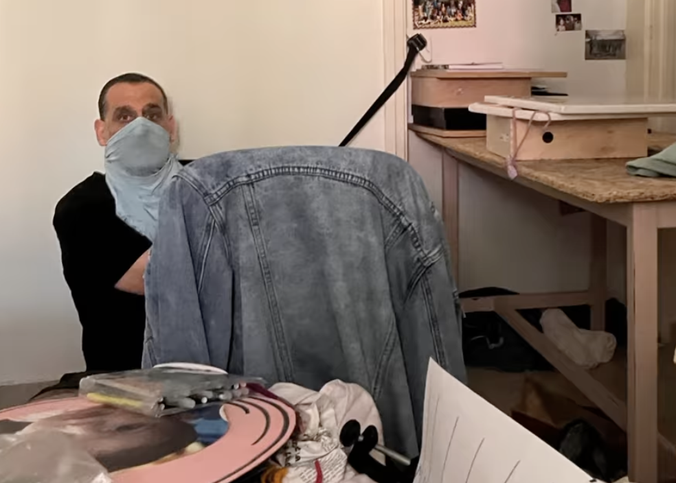 Ein Mann sitzt mit einem T-Shirt über das Gesicht gezogen auf dem Boden eines Zimmers und hält einen Gürtel, der an einer Türklinke befestigt ist