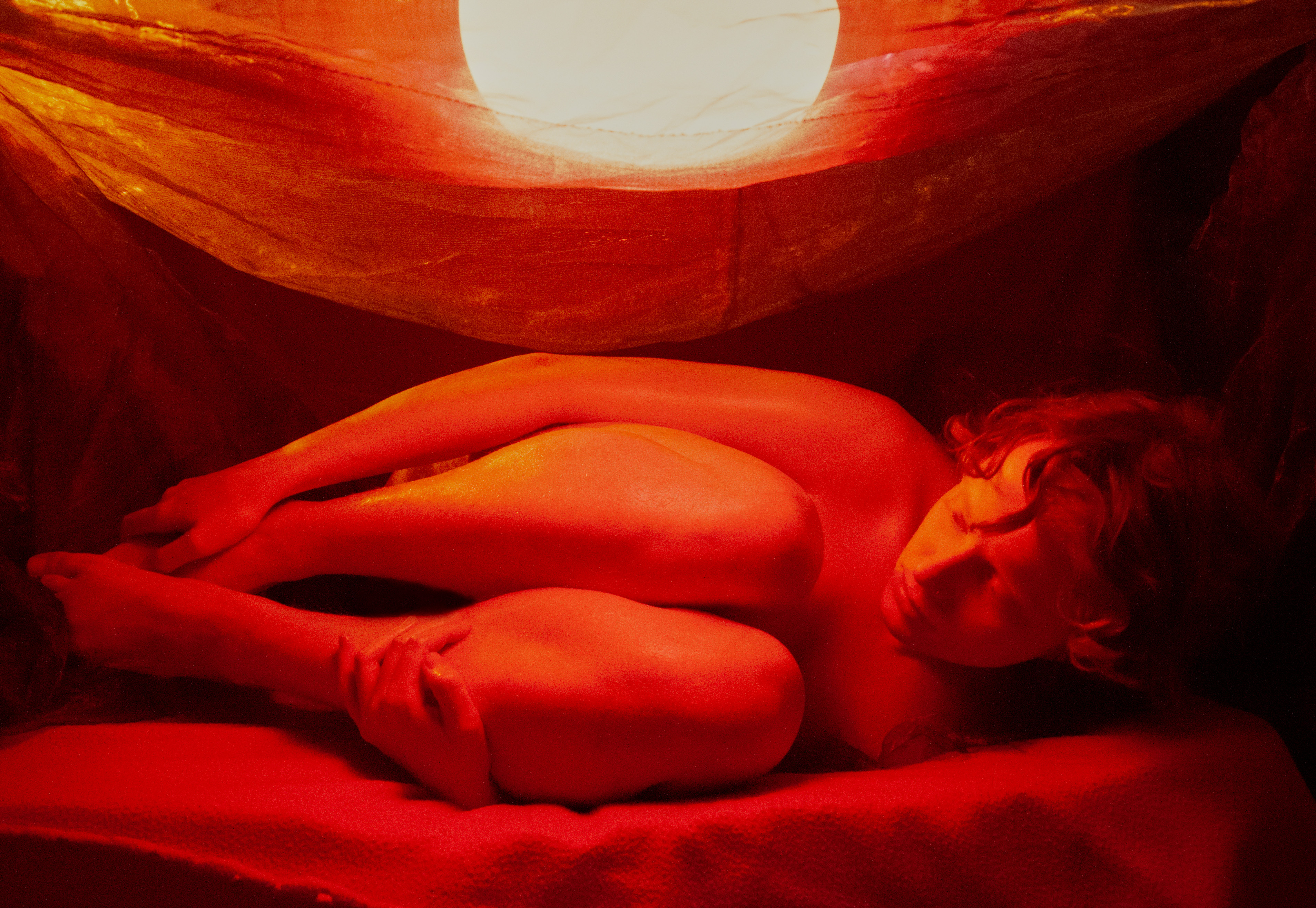 Flo Verhulst – image of a young person in fetal position surrounded by blankets, lit by a red light