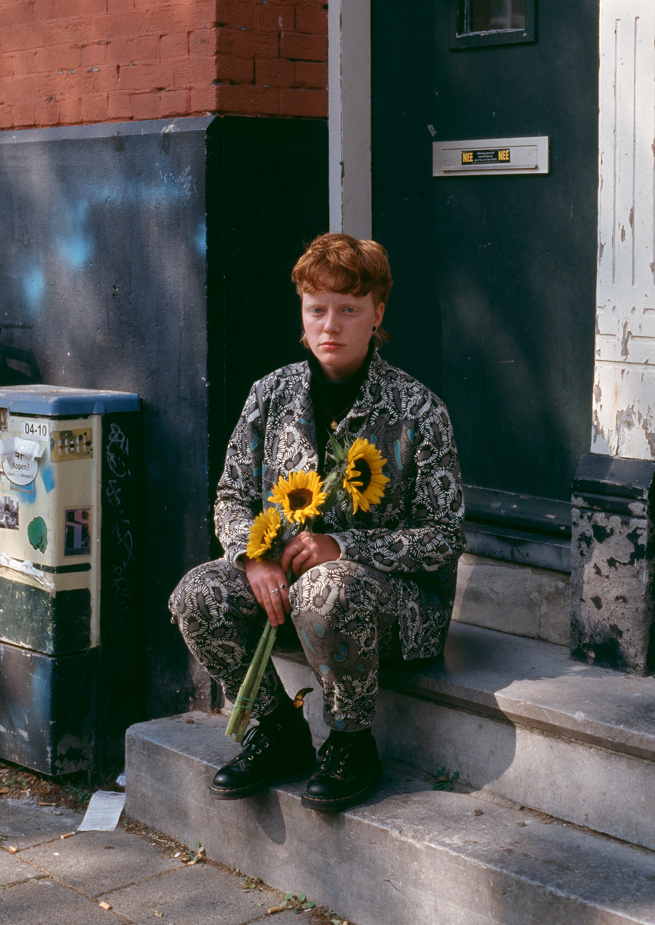 Flo Verhulst – young person with shaggy ginger hair, a funky patterned suit and doc martens, sitting on the stairs in front of an apartment building with three sunflowers in their hand. 