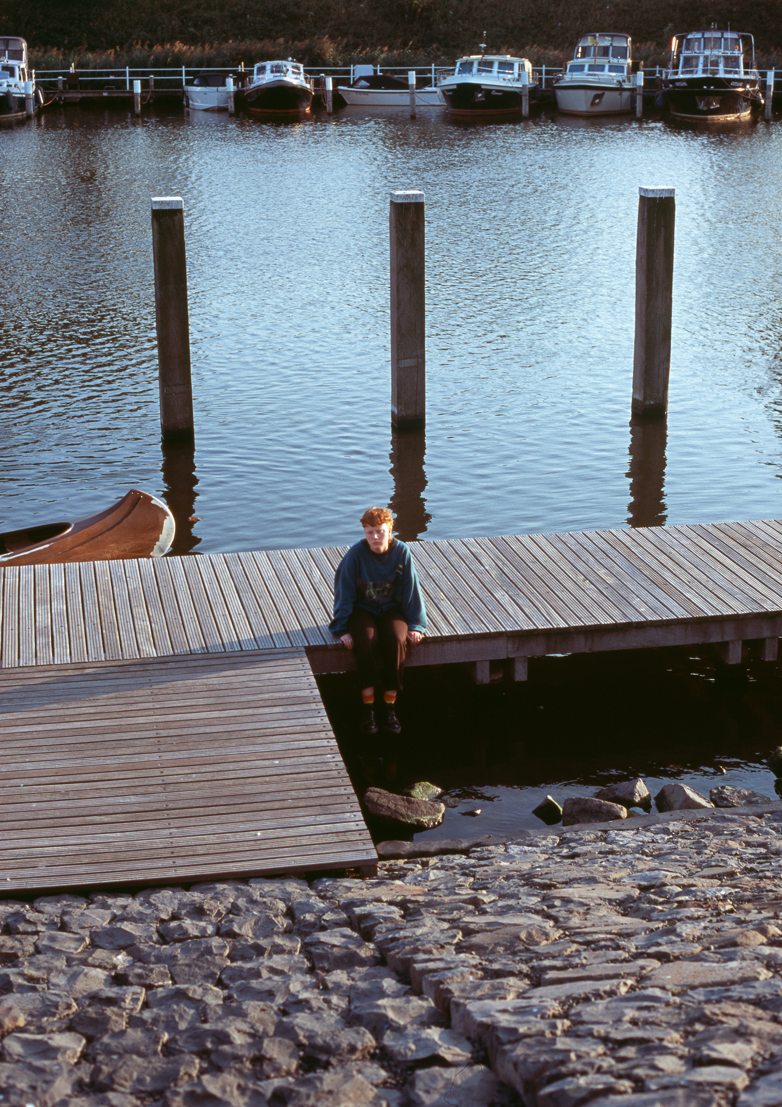 Flo Verhulst – young person sitting on a pier looking sadly to the ground. In the background: water and a few boats