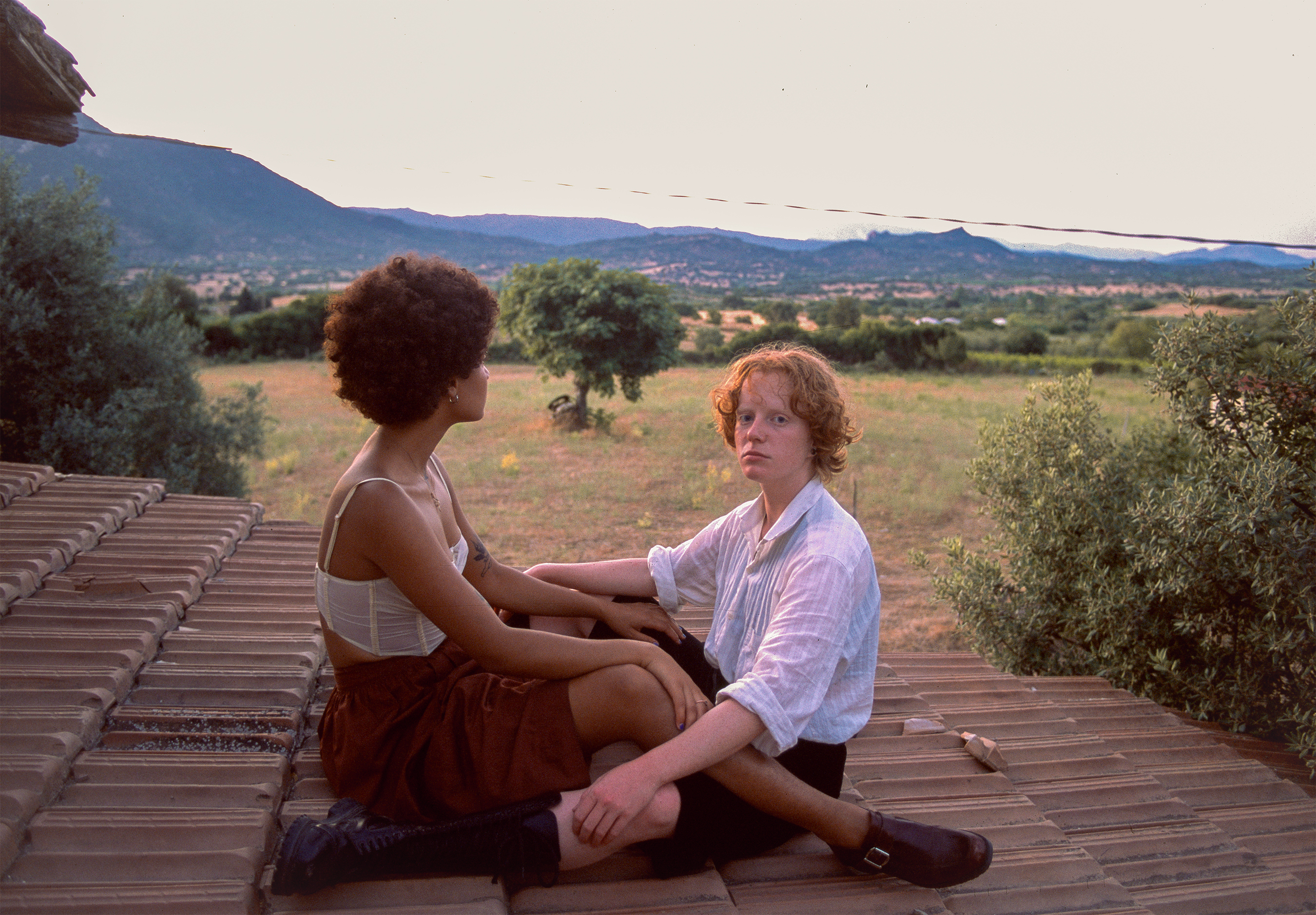 Flo Verhulst – two young people sitting face to face with their legs embracing on top of a roof. One of them is wearing a corset top and a skirt, the other a white shirt, shorts and long socks. Background: countryside