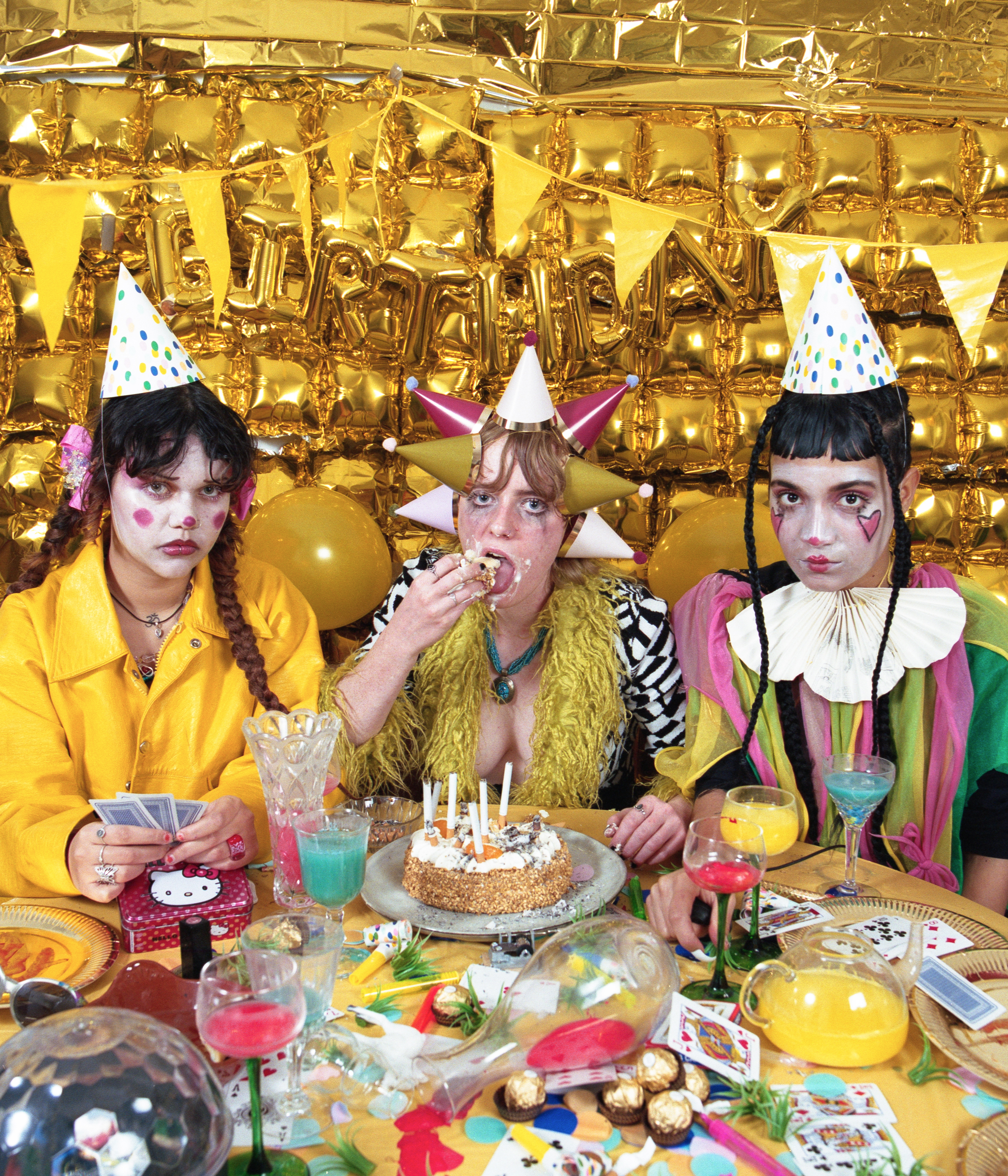 Flo Verhulst – three young people  sitting a table full of stuff, wearing party hats and clown makeup with decorations everywhere in the background. In the middle, flo is eating cake