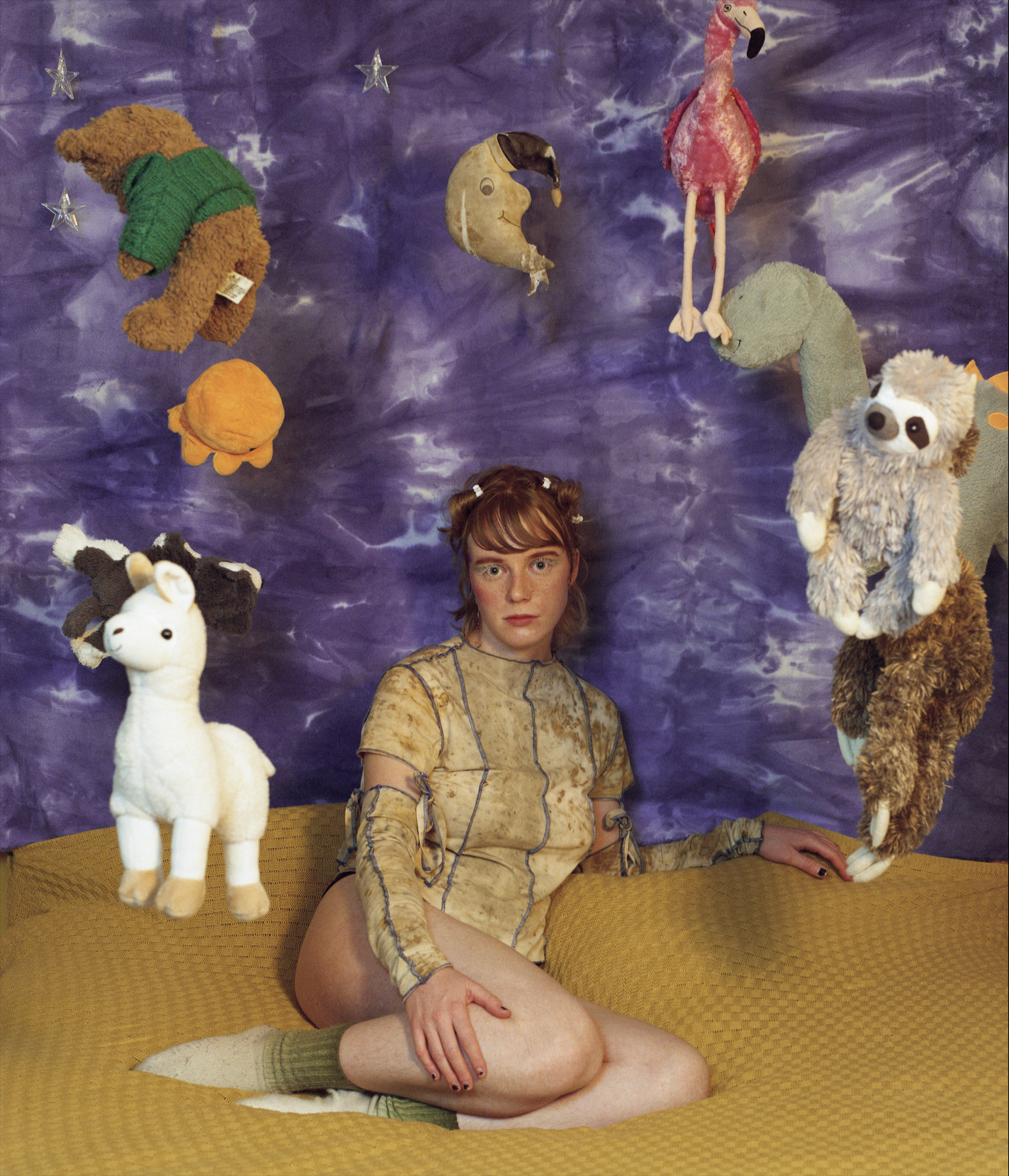 Flo Verhulst – young person wearing a Y2K inspired top, underwear and socks and sitting on a bed, surrounded by stuffed animals floating in the air. Background: a blue tie-dye sheet