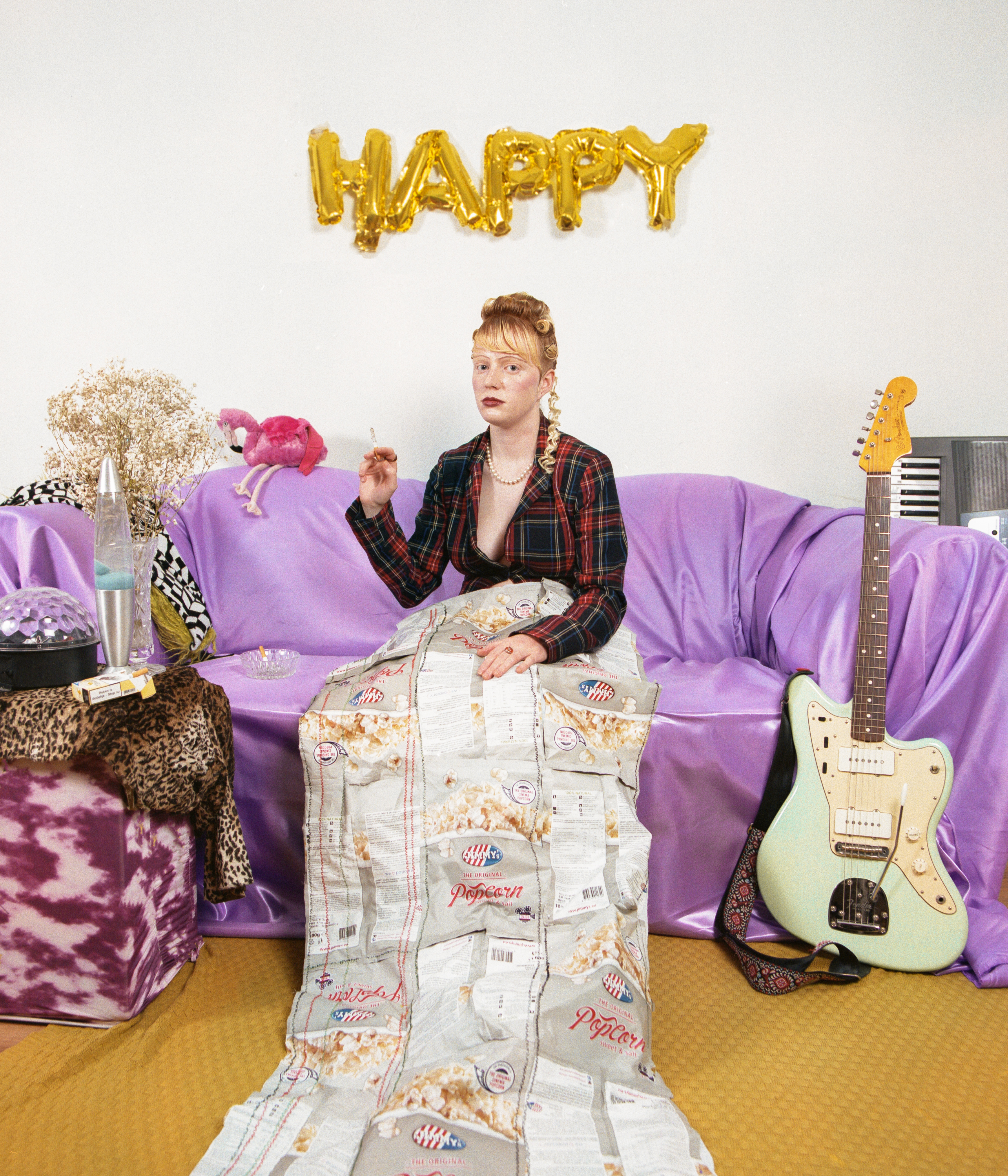 Flo Verhulst – young person in pijamas and an elaborate hairdo, sitting on a purple couch with various objects, smoking a cigarette and looking deadpan at camera