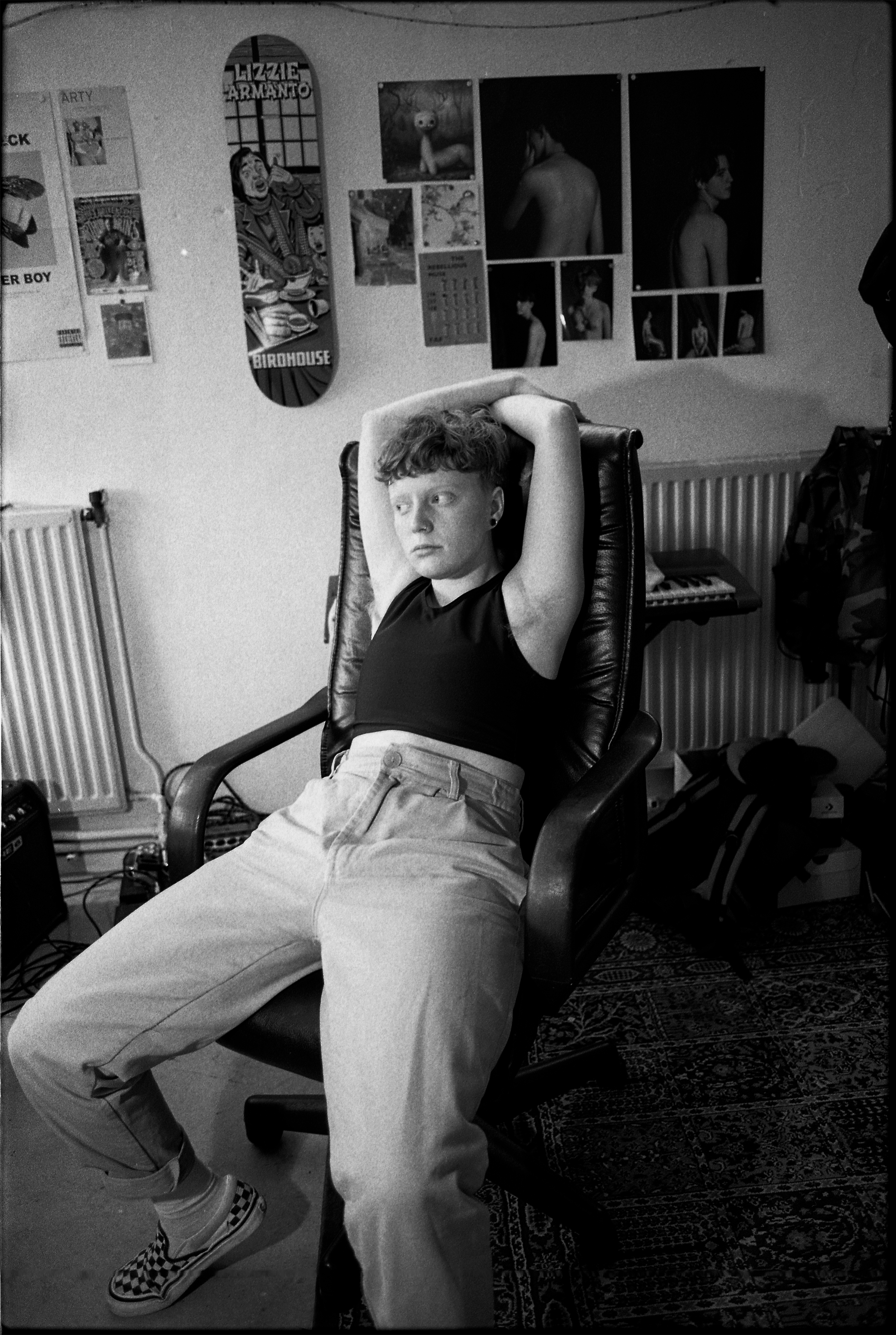 Flo Verhulst – young person wearing a black crop top, cuffed jeans and checkered vans slouching on a desk chair with their arms crossed above their head and looking into the distance