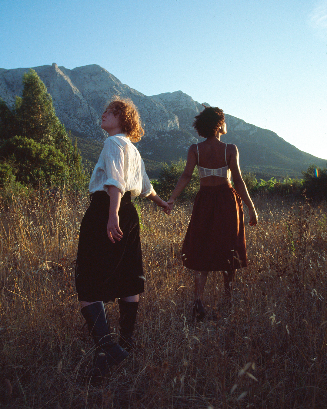 Flo Verhulst - photo of two people holding hands in a sunny wheat field with a mountain in the background. One is ginger and wears a white shirt with black skirt, the other has dark, curly har, a brown skirt and a white bra