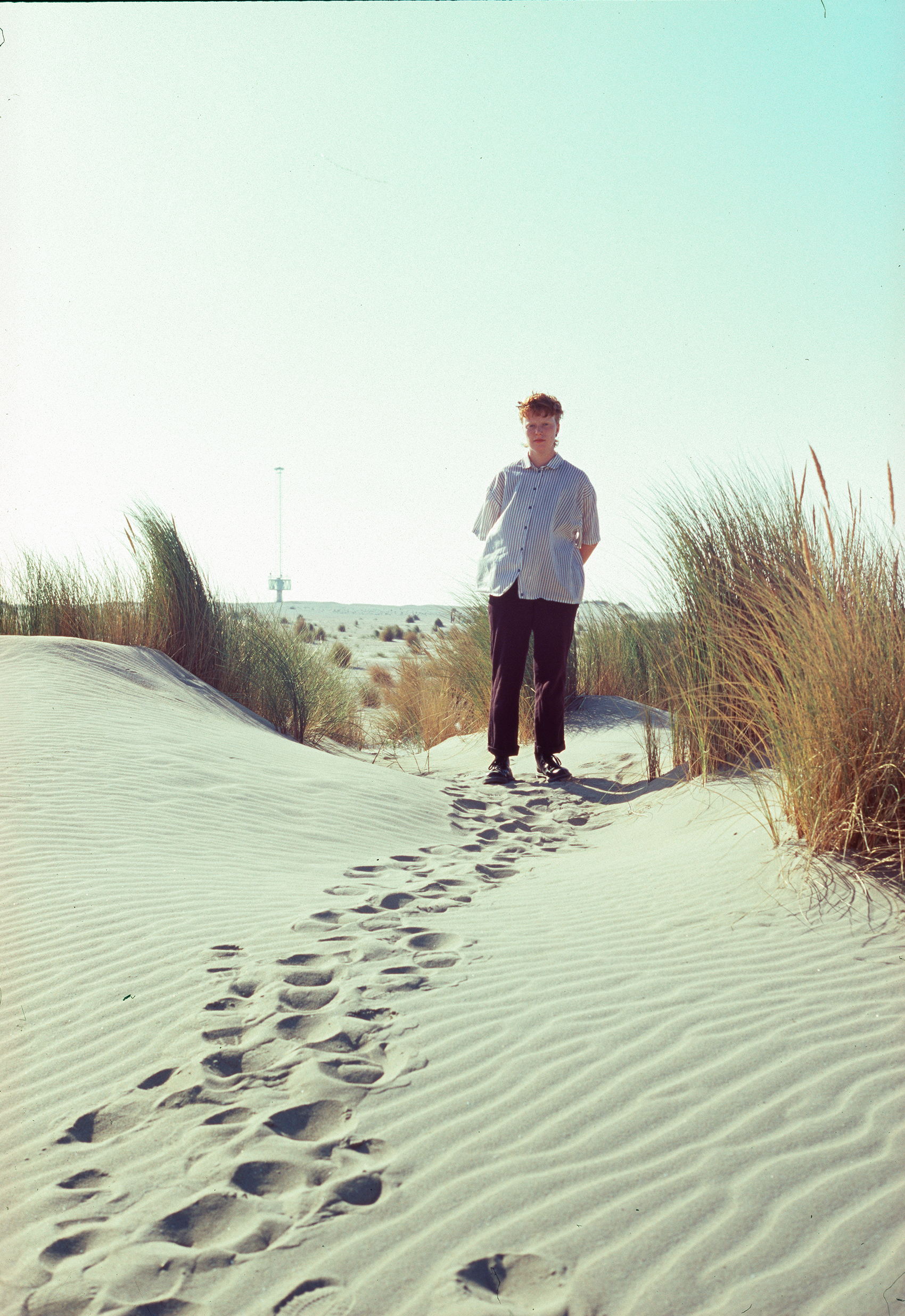 Flo Verhulst - photo of a ginger person standing on sand dunes with tall grass. They wear black pants and a white and blue shirt.