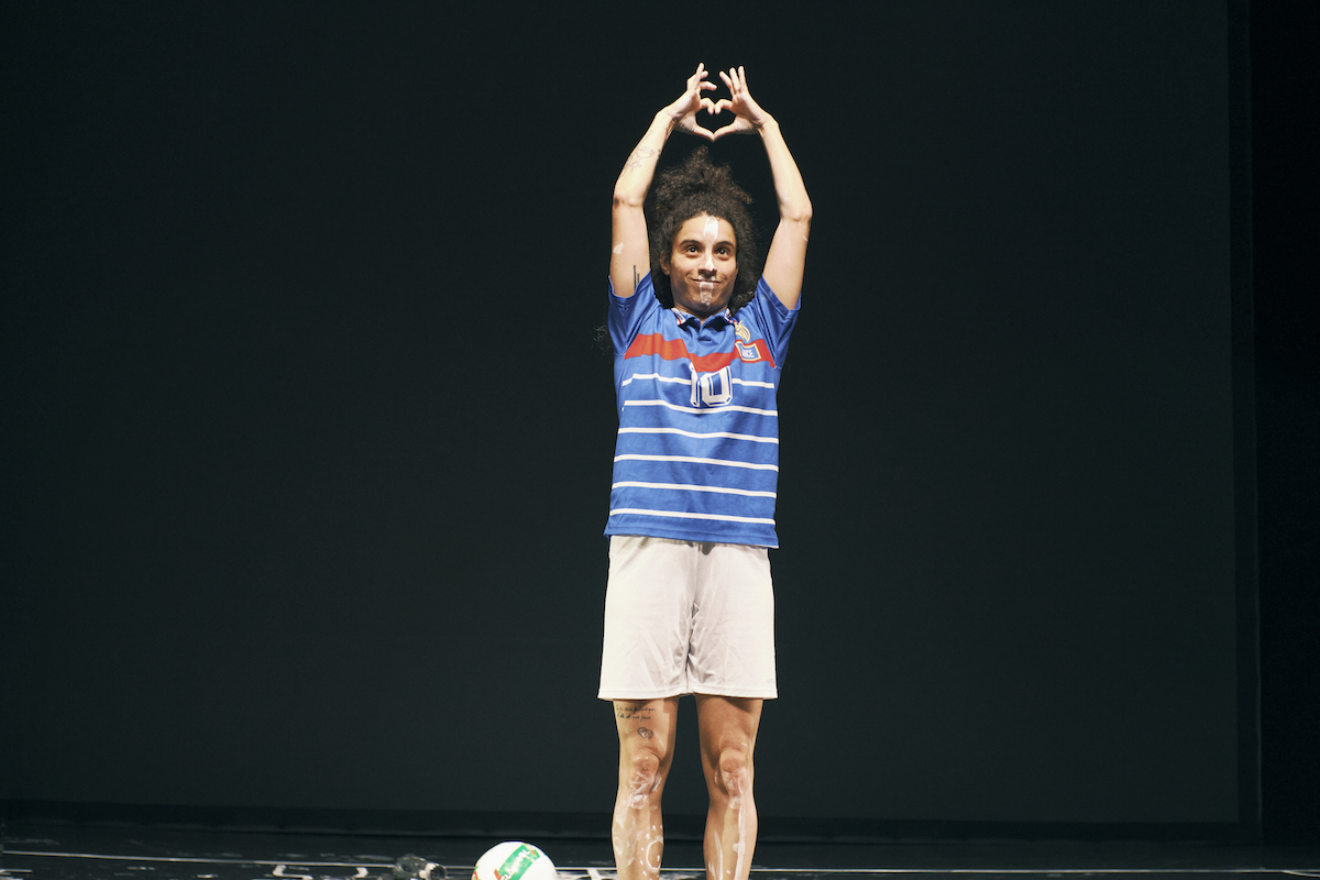 photo of a performer on stage wearing a blue football jersey and white peint on her skin. There's a ball on the floor and she makes a heart with her two hands