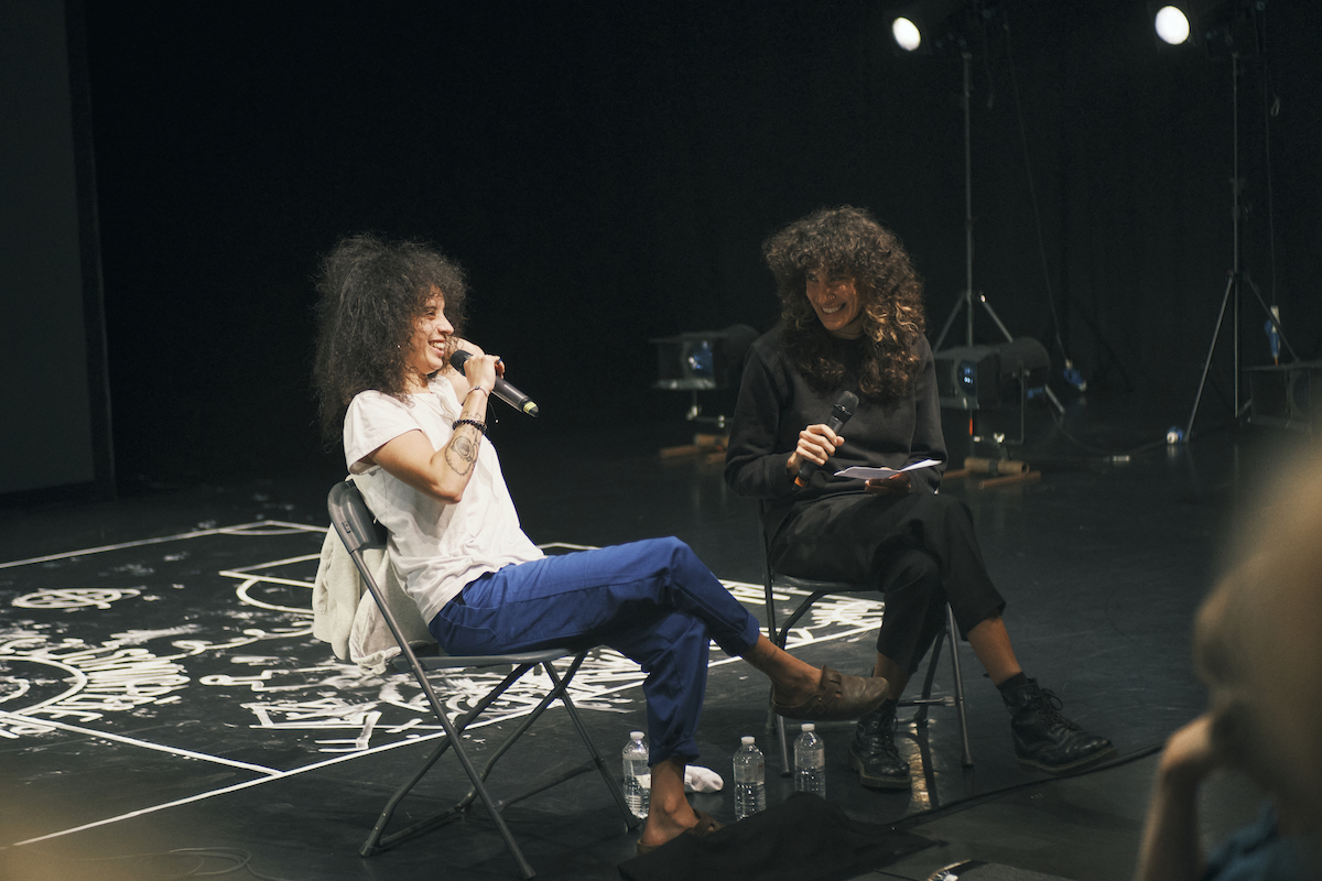 Two women with dark curly hair having a chat on stage, they both hold a microphone and there's a white drawing on the floor in the background. One is dressed in black and the other is wearing a white t-shirt and blue pants