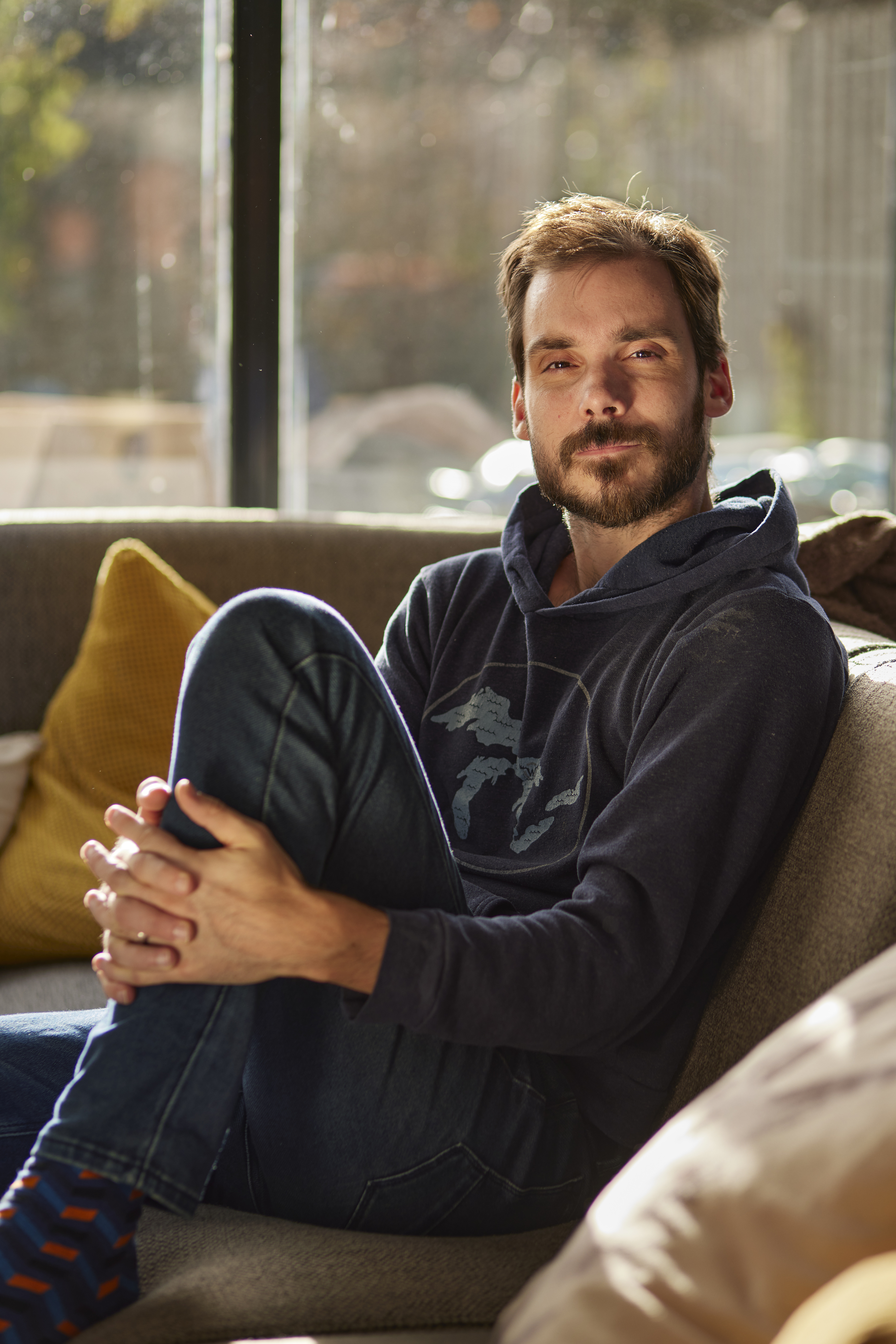 portrait of a white bearded man wearing blue jeans and a dark blue hoodie, he's sitting on his couch, holding one of legs and looking at the camera. There's a big window in the background