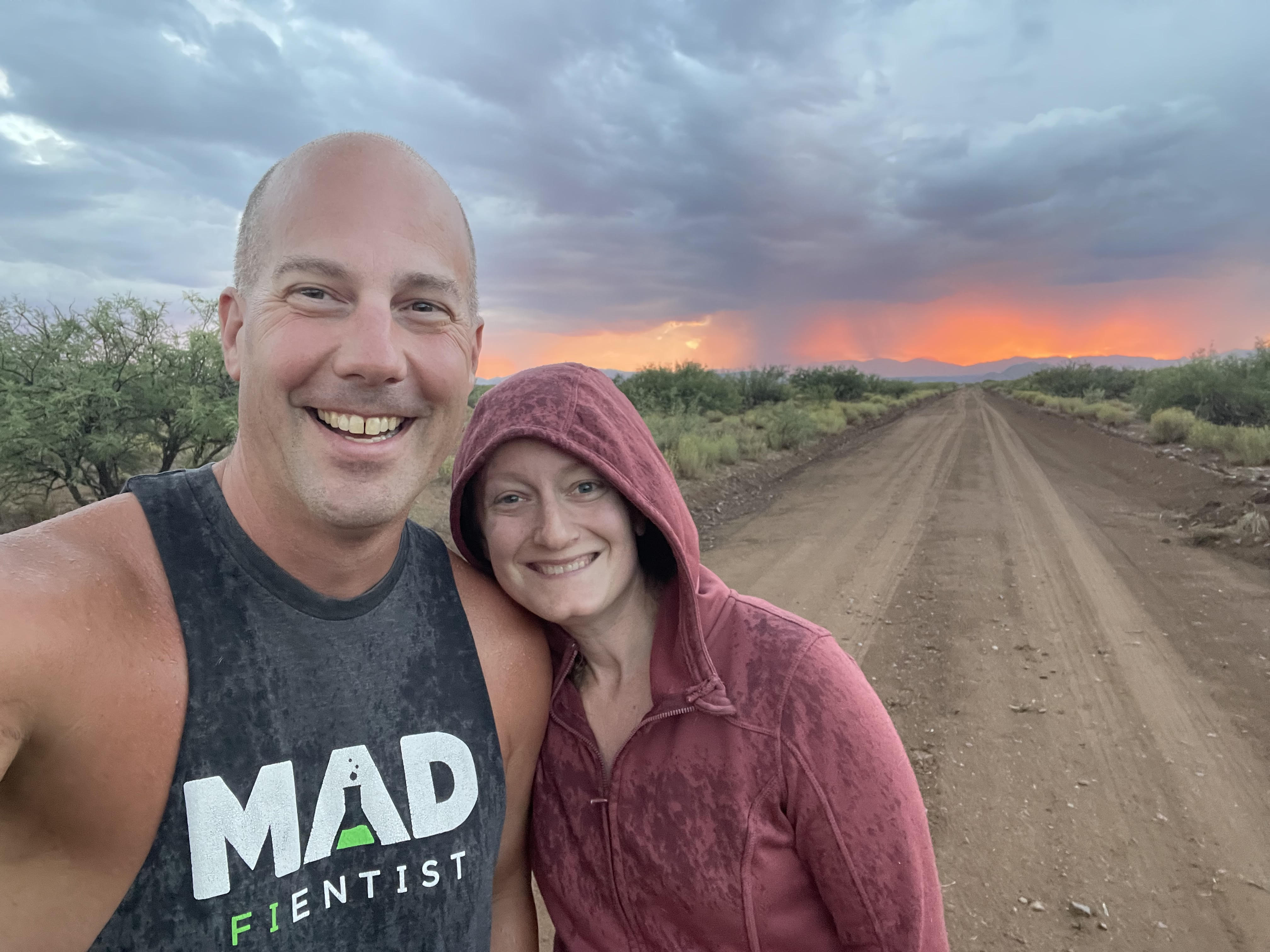 A man and his wife pose for a photo next to a dirt road with a orange sunset behind them.