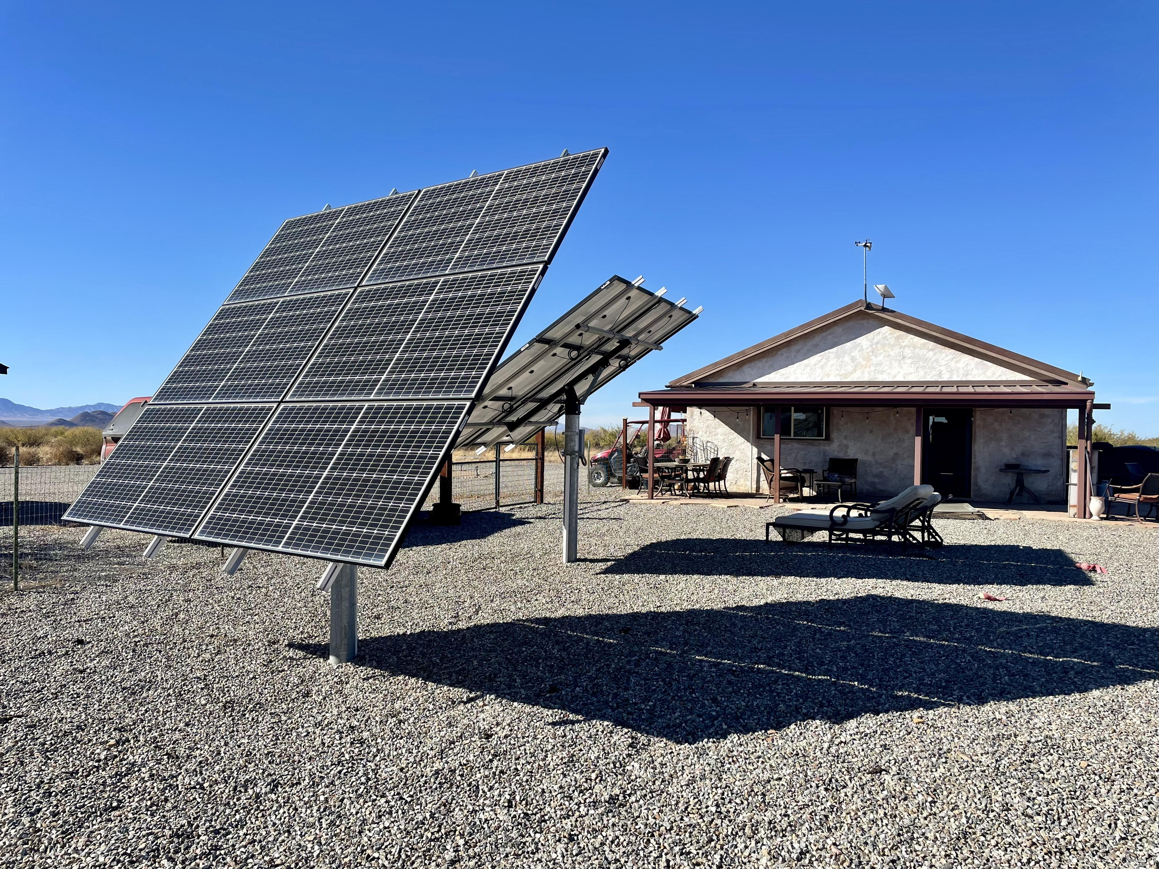 A photo of a one storey house in the desert with two large solar panels in the front yard.