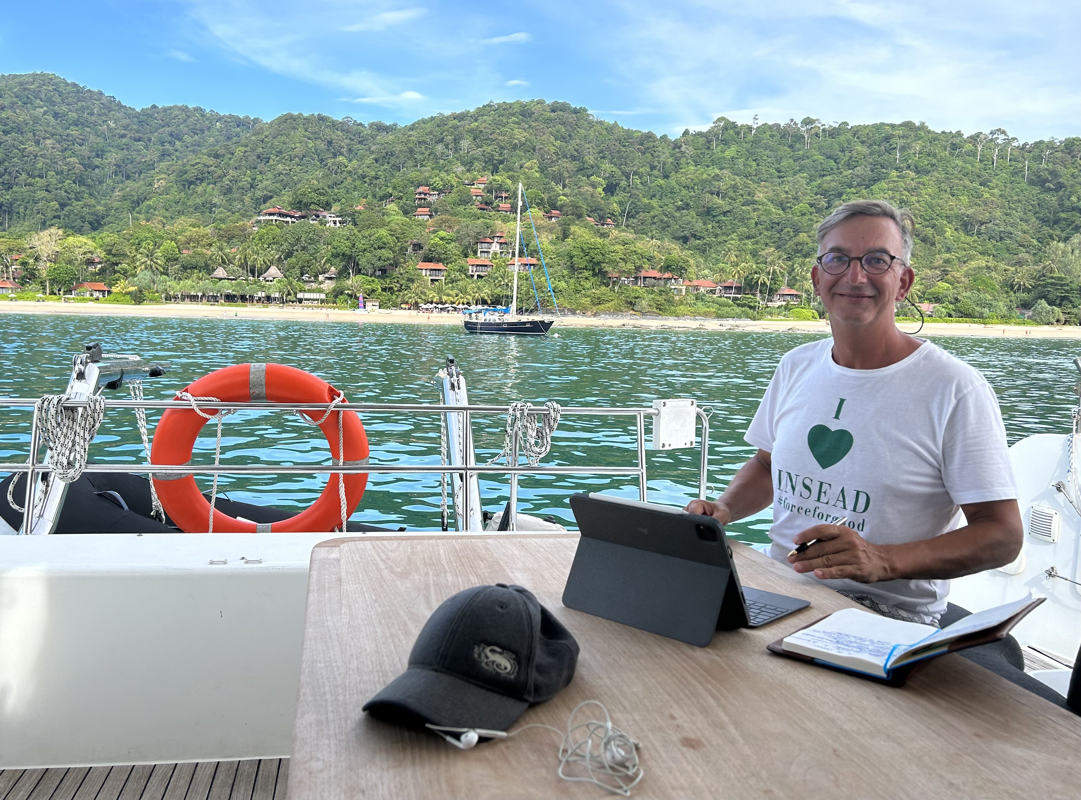 A man on a boat uses his laptop and poses for the camera; there is a picturesque island in the background.