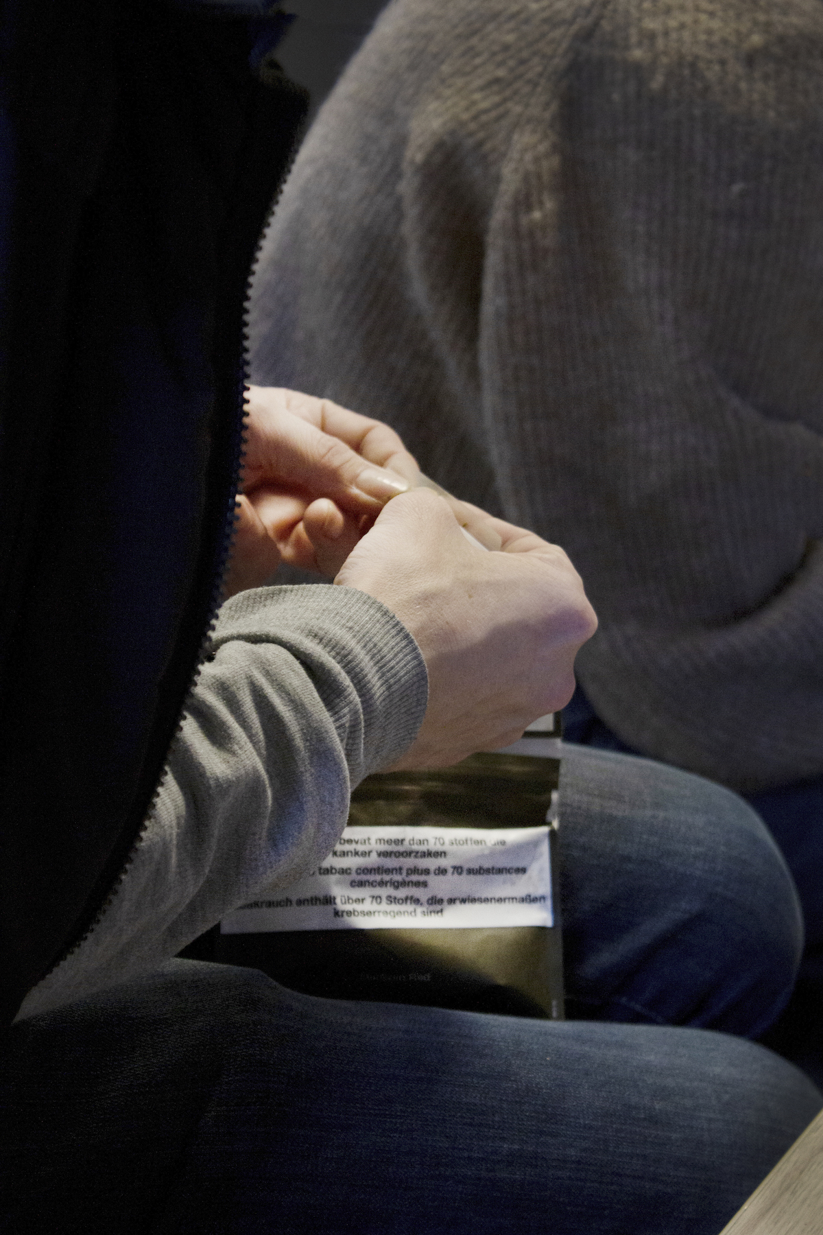 Transit, Brussels – closeup of a man's hands rolling a cigarette. He is wearing a grey sweatshirt and blue jeans.