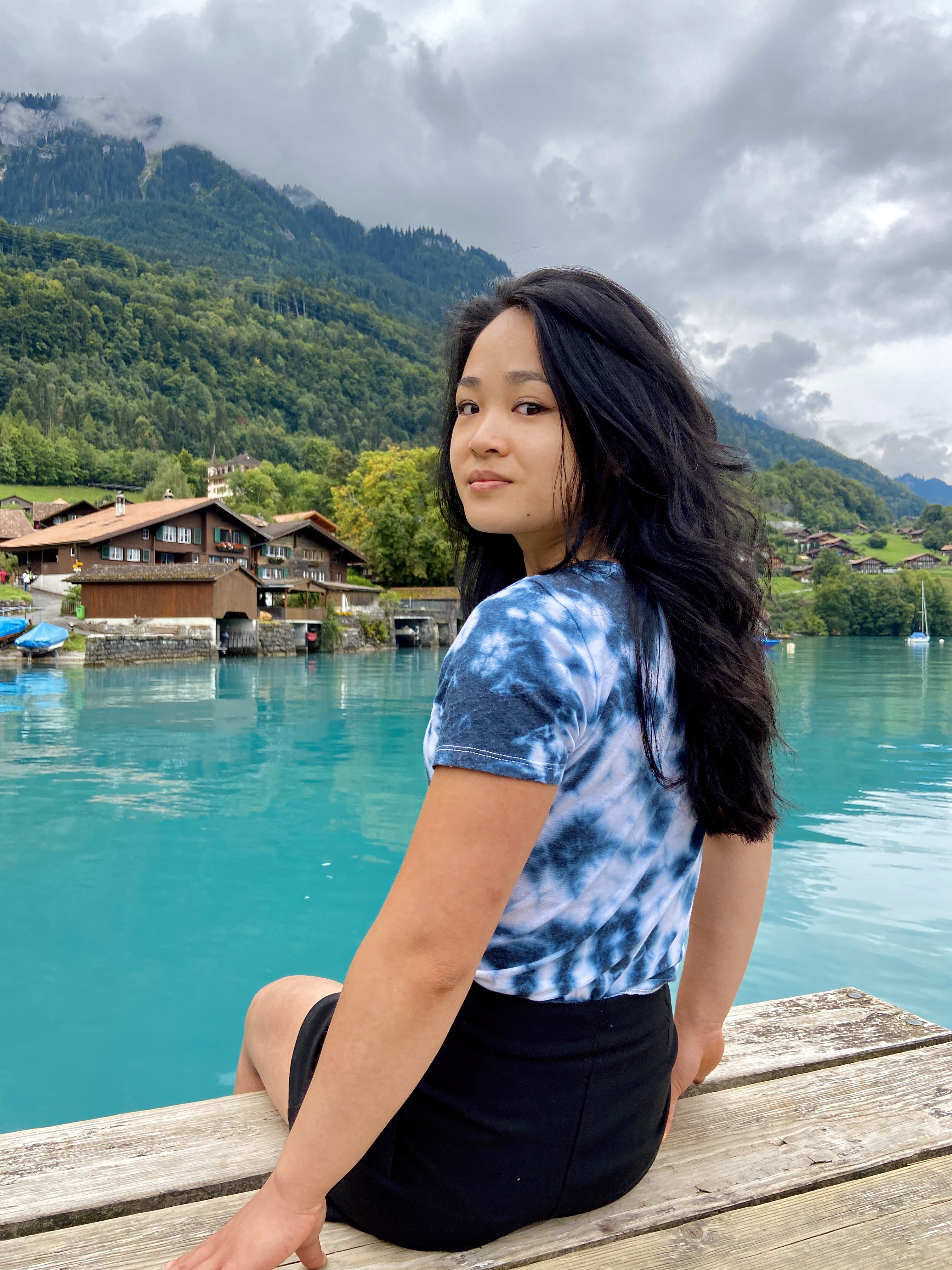 Xiangxia van den Ham, Dumpling Stories – young woman wearing a tie die t-shirt and a tight black skirt sitting by a turquois lake surrounded by wooden houses. 