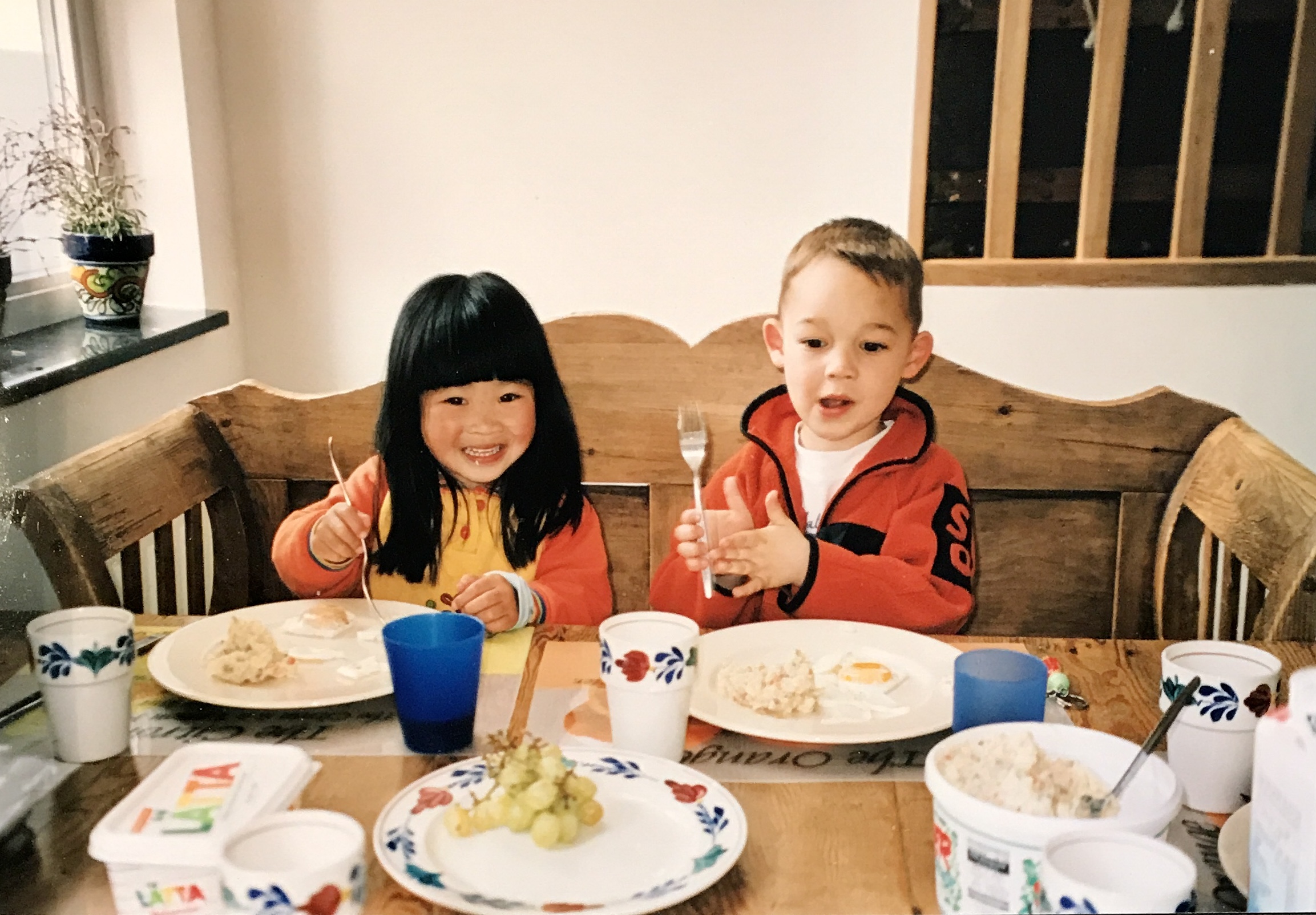 Xiangxia van den Ham, Dumpling Stories – a boy and a girl sitting side by side at a large wooden table with white and blue plats and mugs and having breakfast. Xiangxia (left) has long black hair and a fringe and smiling big.