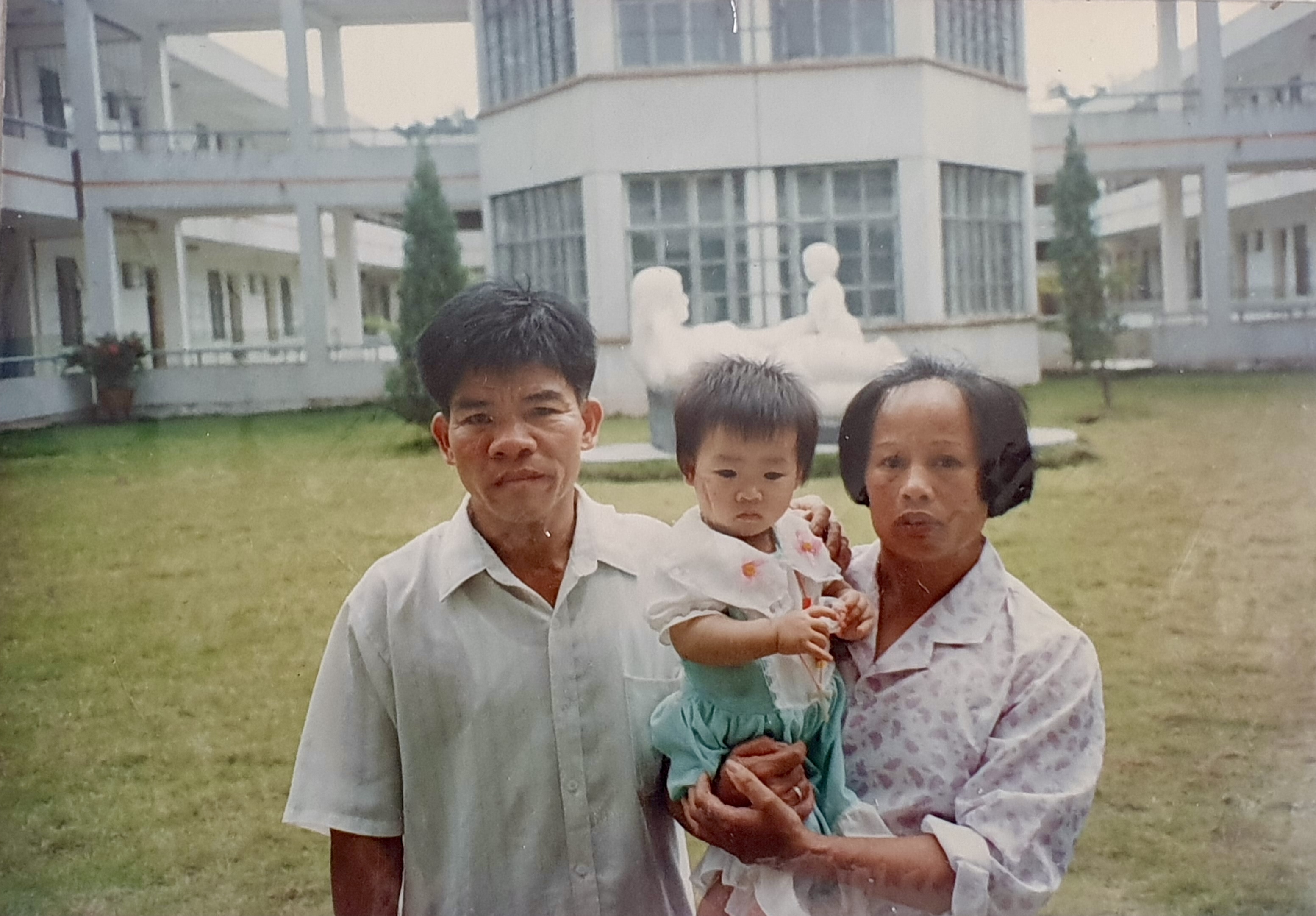 Xiangxia van den Ham, Dumpling Stories – middle aged couple holding a small child in front of a large white building with a statue of a mother and a baby