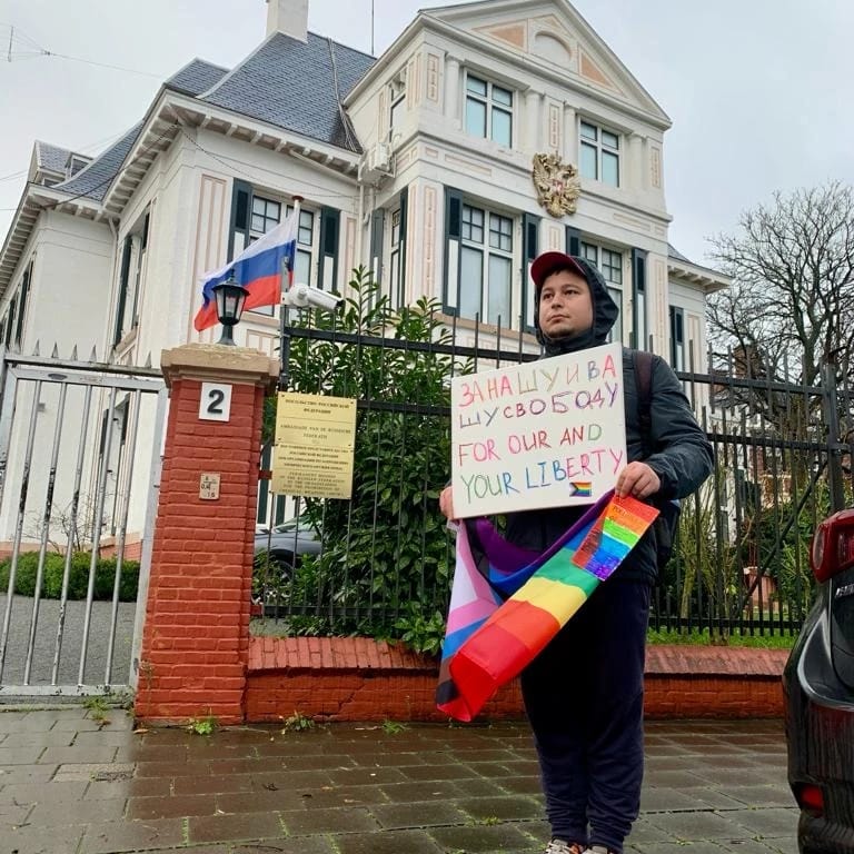 Ein Mann steht mit Schild und Regenbogenflagge vor einem Haus, vor dem die russische Flagge weht