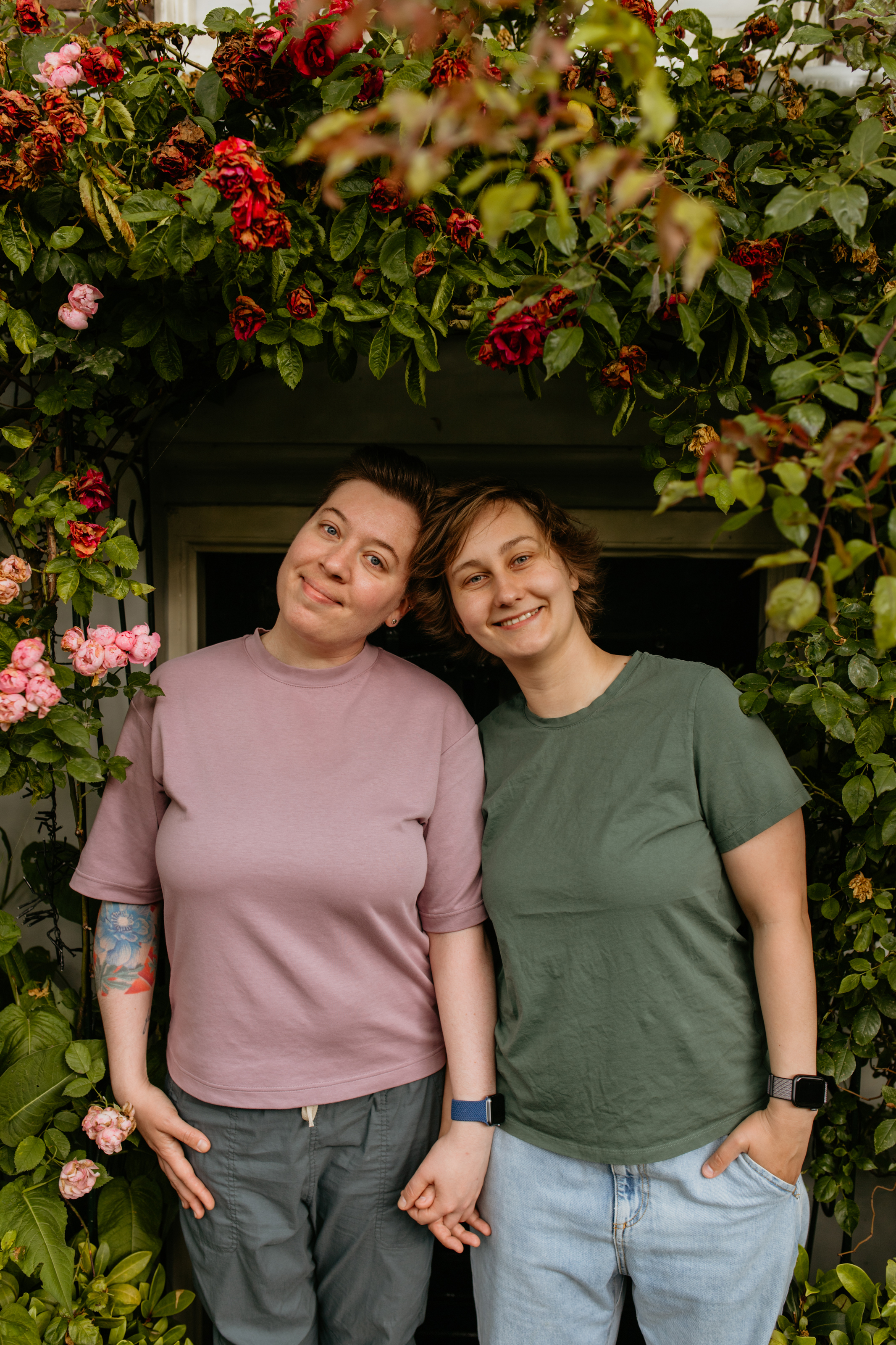 Zwei Frauen mit kurzen Haaren halten zwischen Rosen Händchen und schauen lächelnd in die Kamera