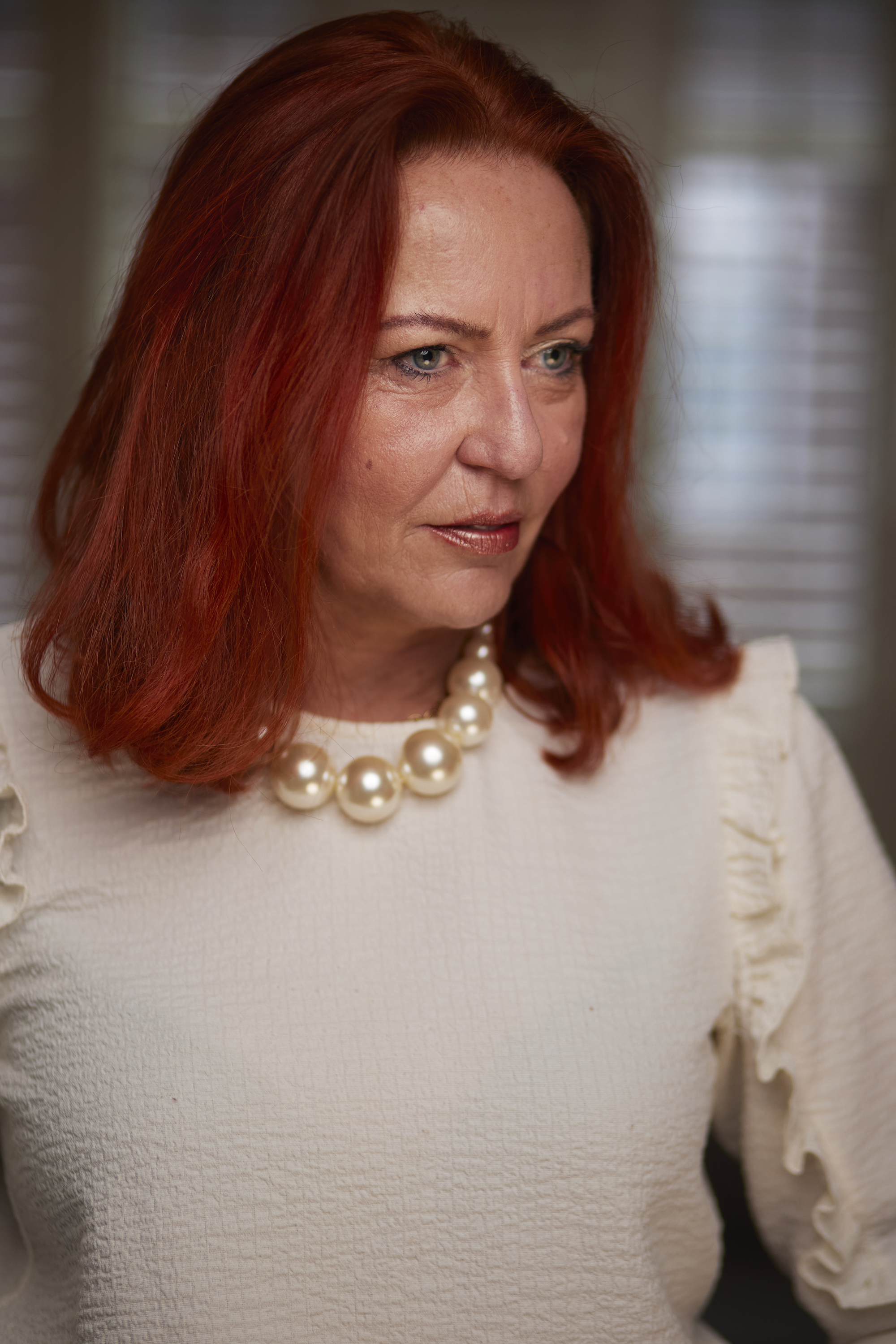 portrait of a woman with long, red hair and blue eyes wearing a big necklace with pearls and a white blouse.