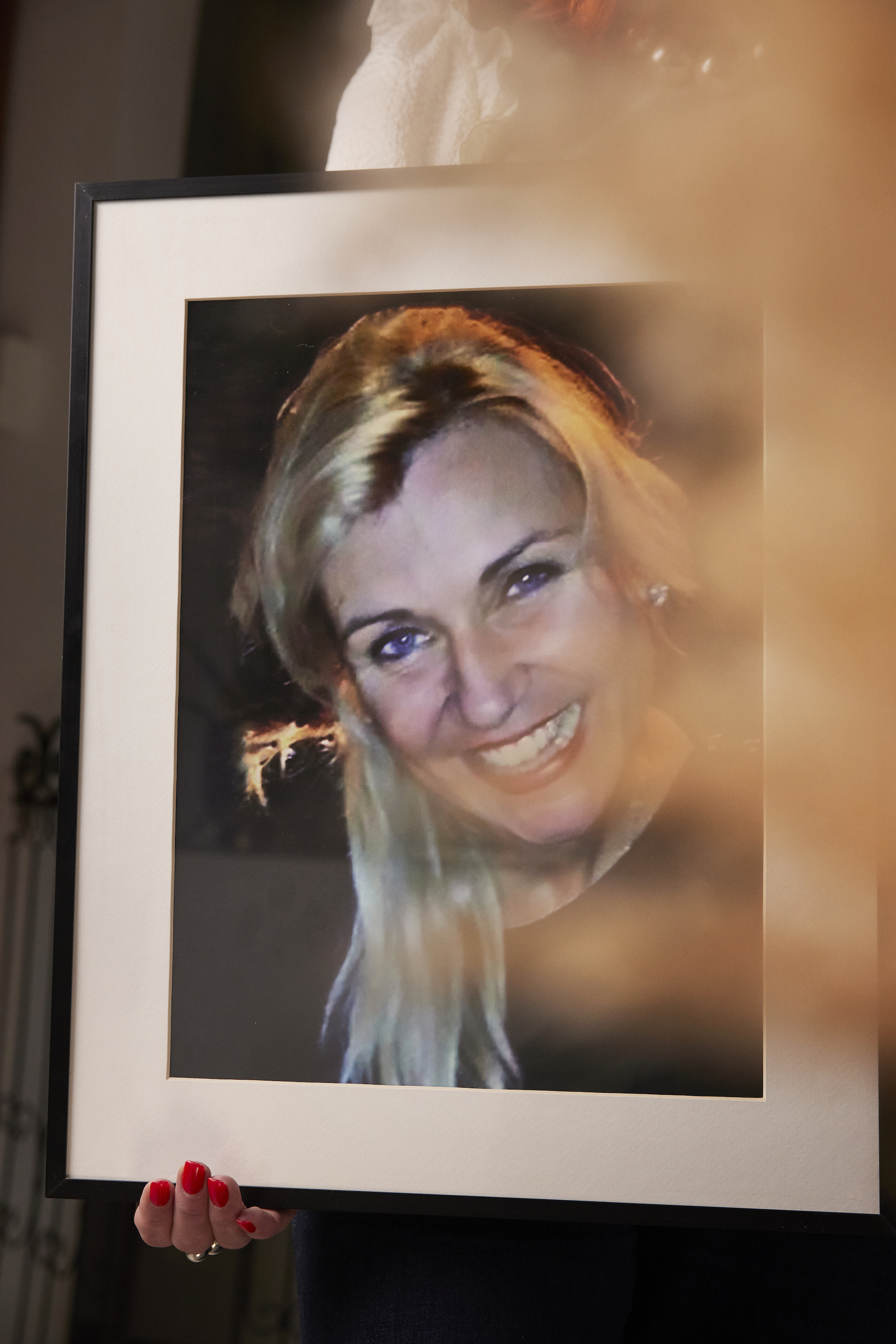 photo of a picture of a blond girl in a frame. The girl has long hair and smiles and the camera, and the frame is held by hands with red nail polish