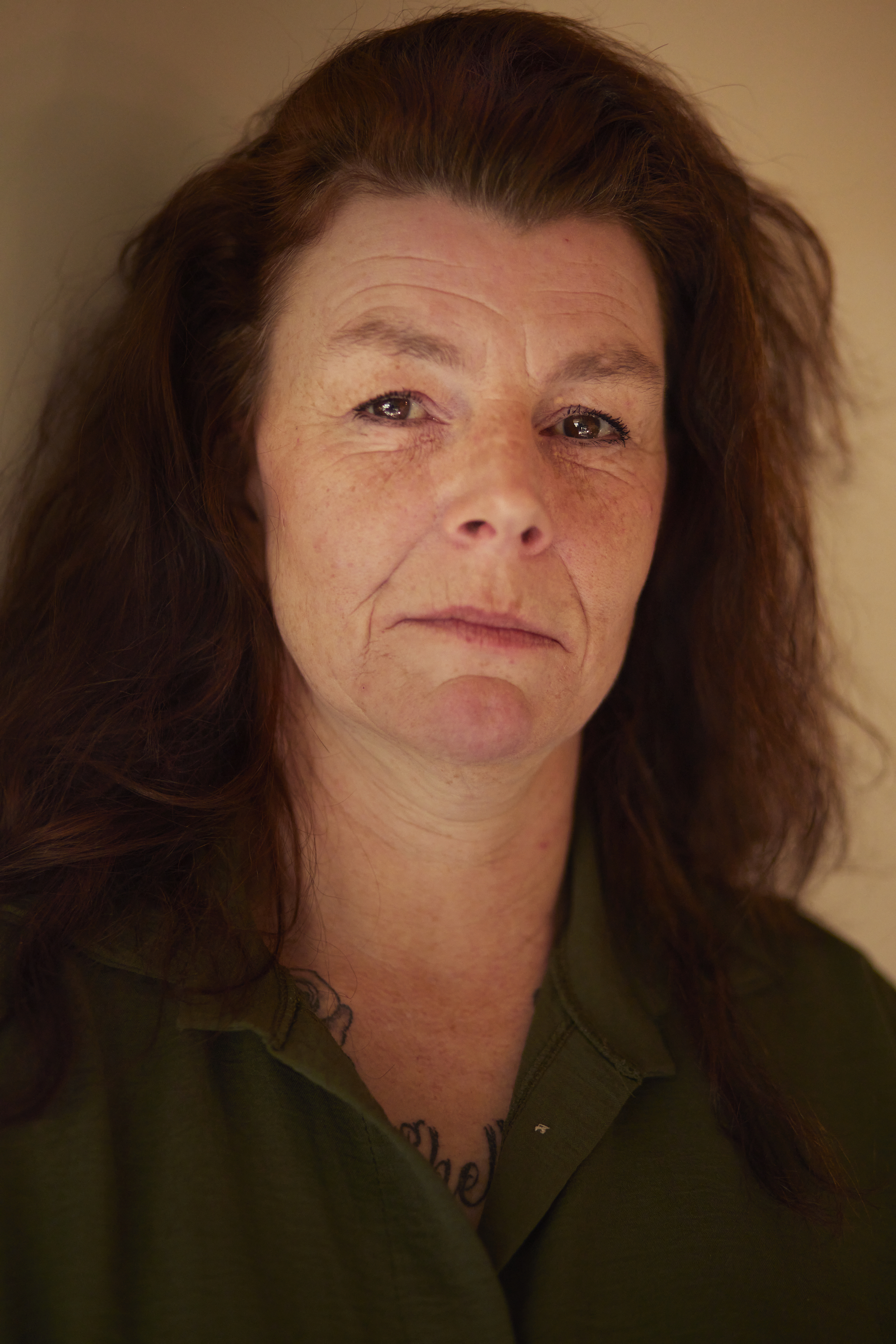 portrait of a woman with long, brown hair wearing a kaki shirt. She has a tattoo on her chest