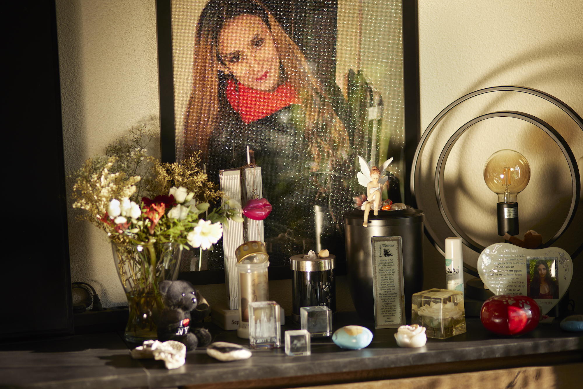 photo of an autel with a big photo of a young woman with long hair, a leather jacket and red jumper. There are flowers, a lamp and several objects in front of the photo