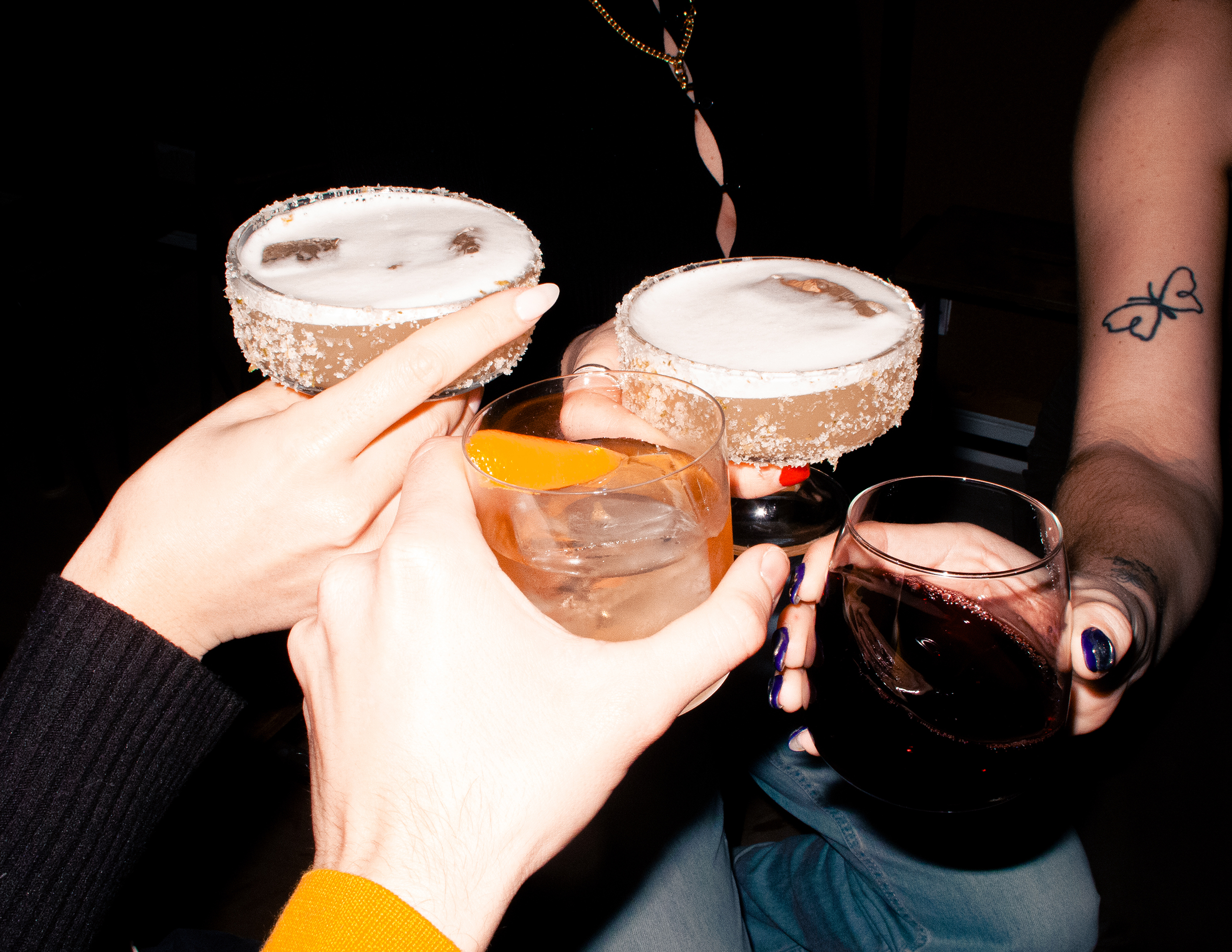 A photo of four arms holding cocktails and cheers-ing in a bar.