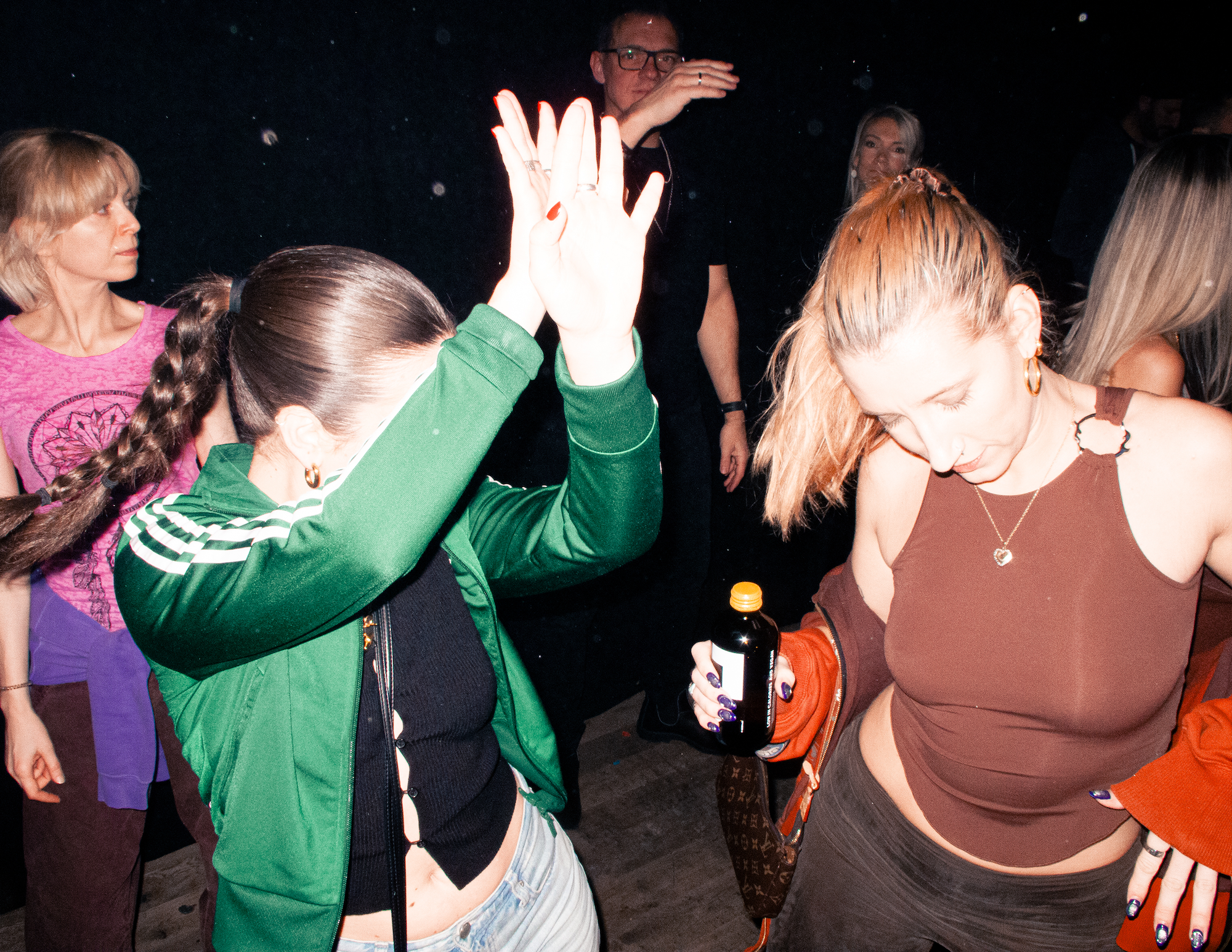 A photo of two girls dancing in a club, one is wearing a green adidas tracksuit top, the other is wearing a brown vest top holding a kombucha bottle.