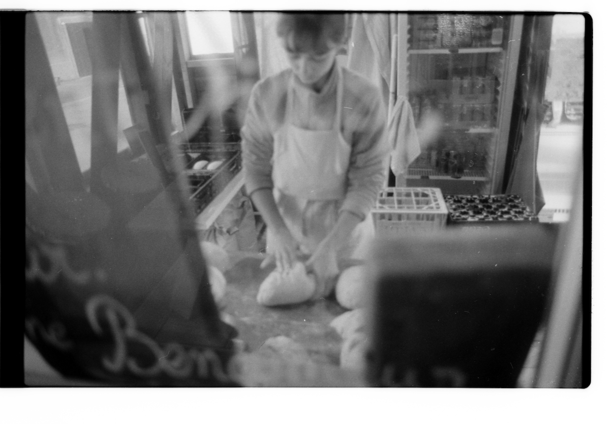 militant bakers - black and white blurry picture of a young woman making bread in a kitchen with a fridge in the background. She's wearing an apron