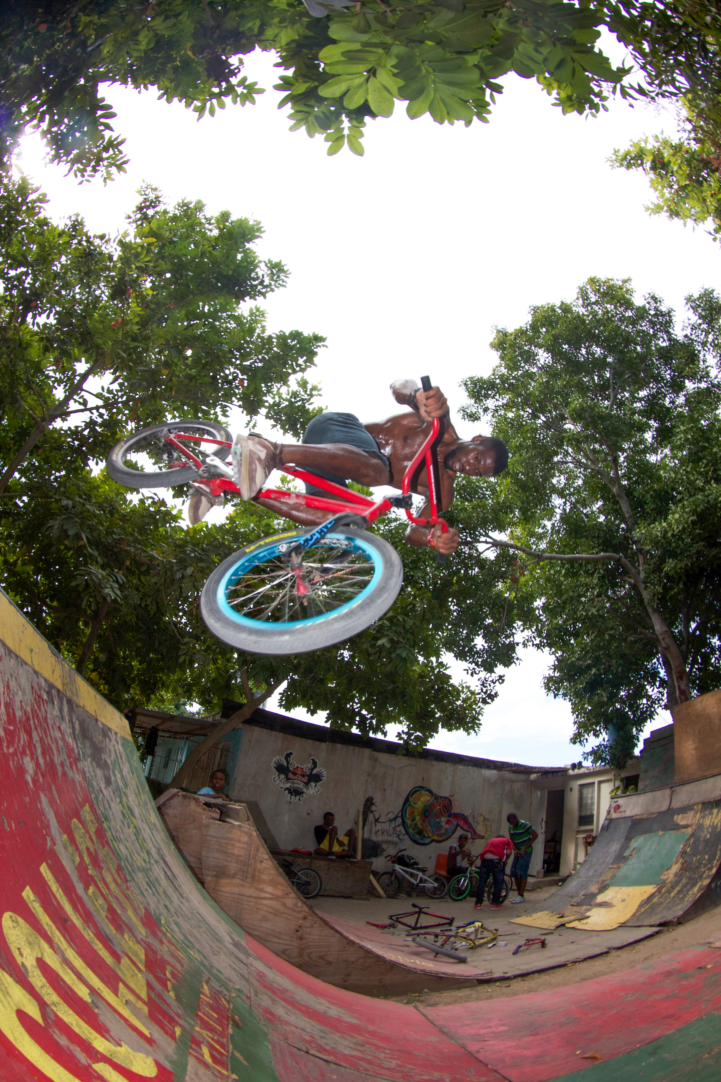 A topless man leaps through the air on a red BMX above a wooden ramp.