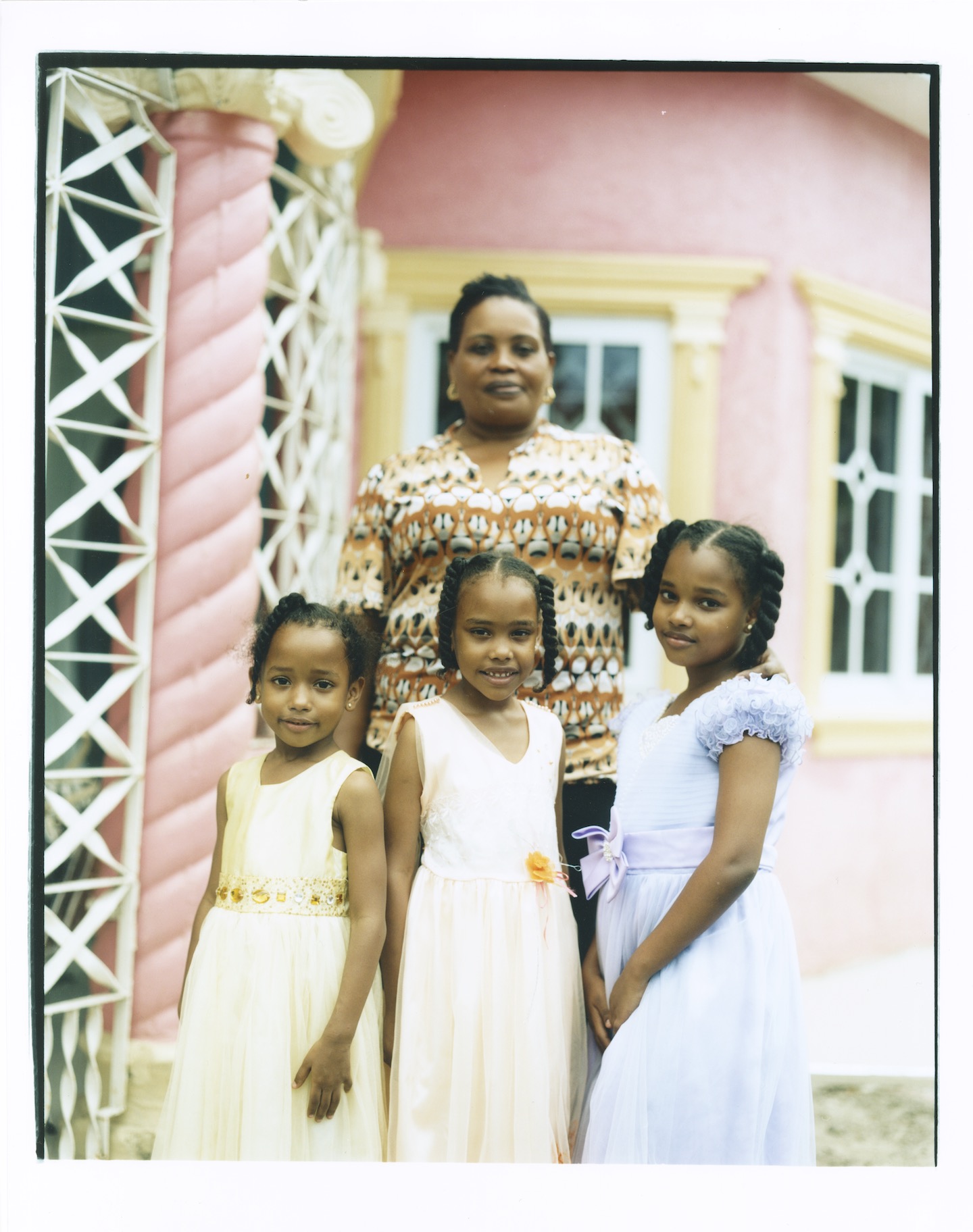 An older woman poses with her three daughters, all wearing dresses, outside a pink house.