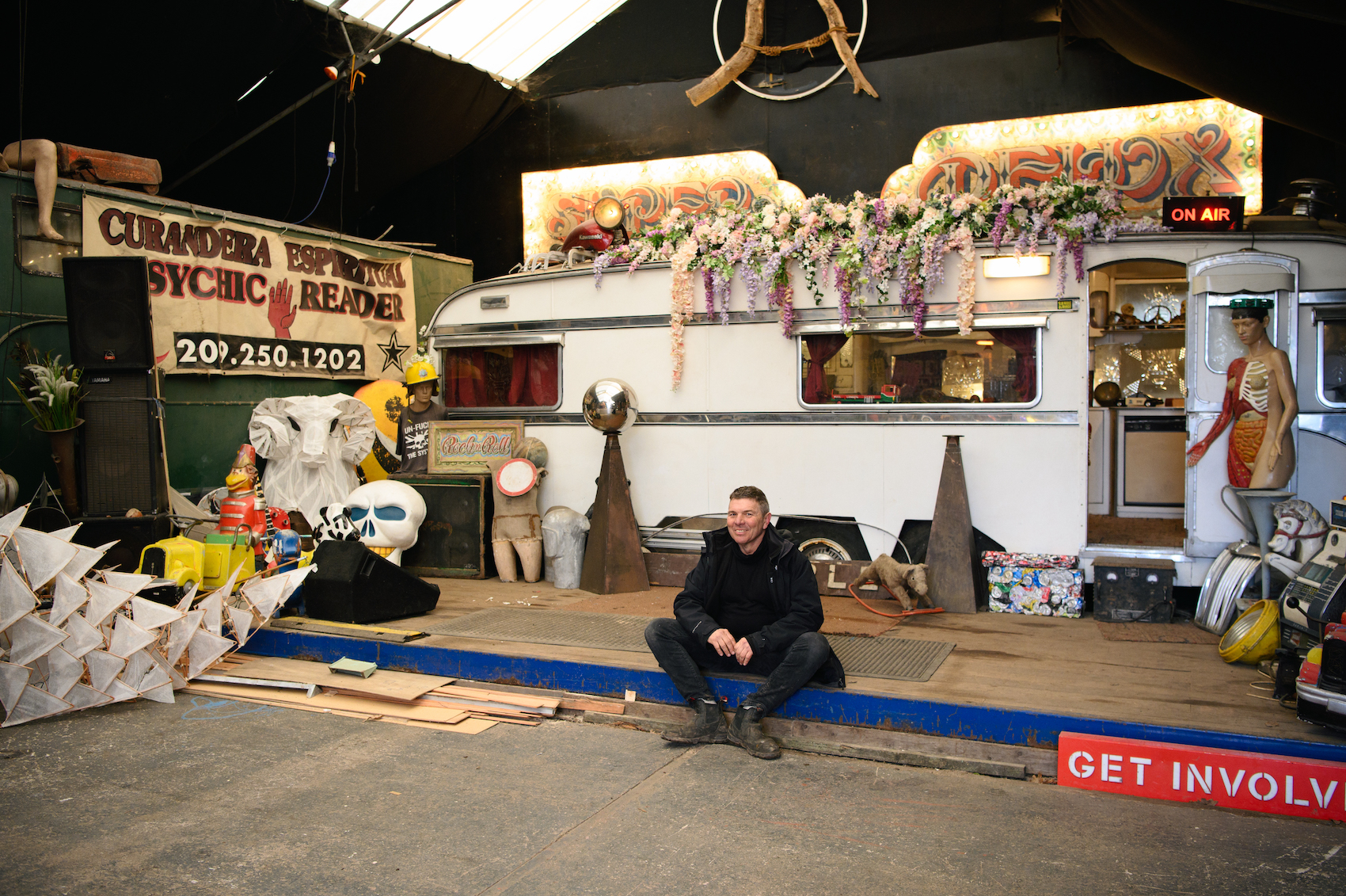 A man sits in infront of a range of eccentric objects and a caravan.