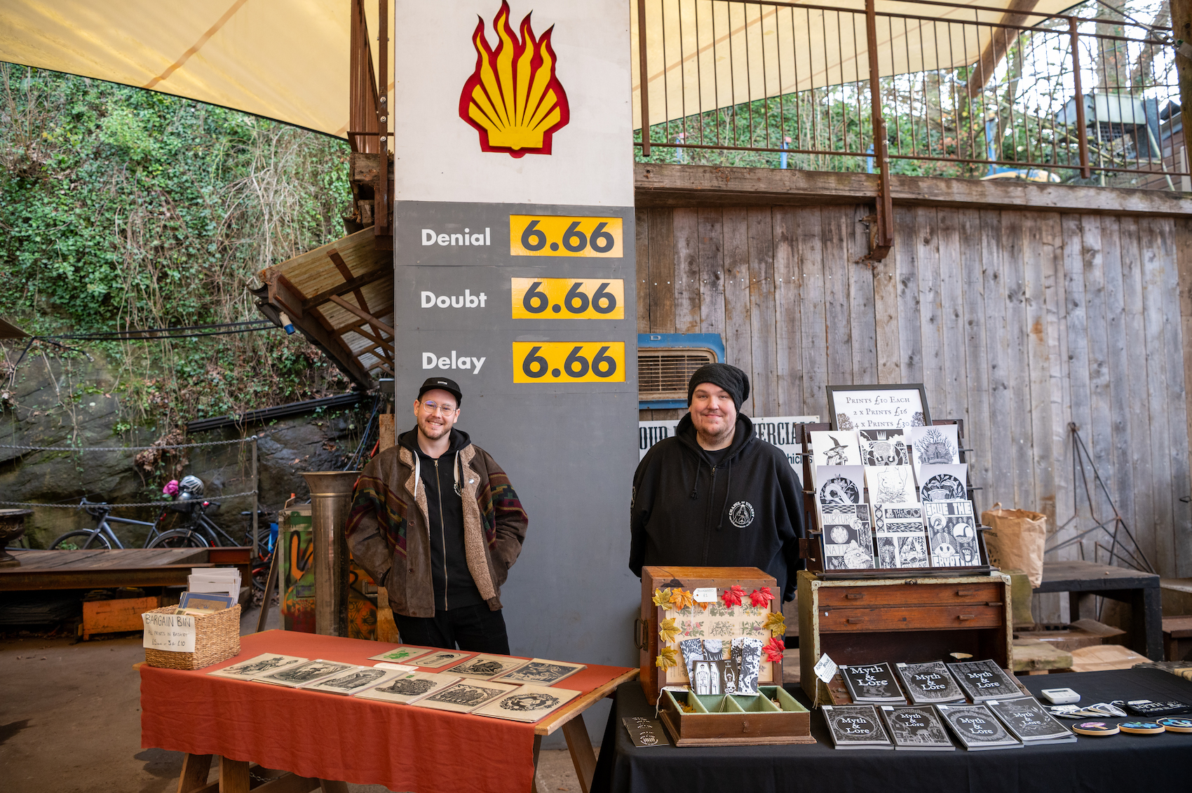 Two men stand at a stall selling zines, behind them is a large sign satirising petrol stations.