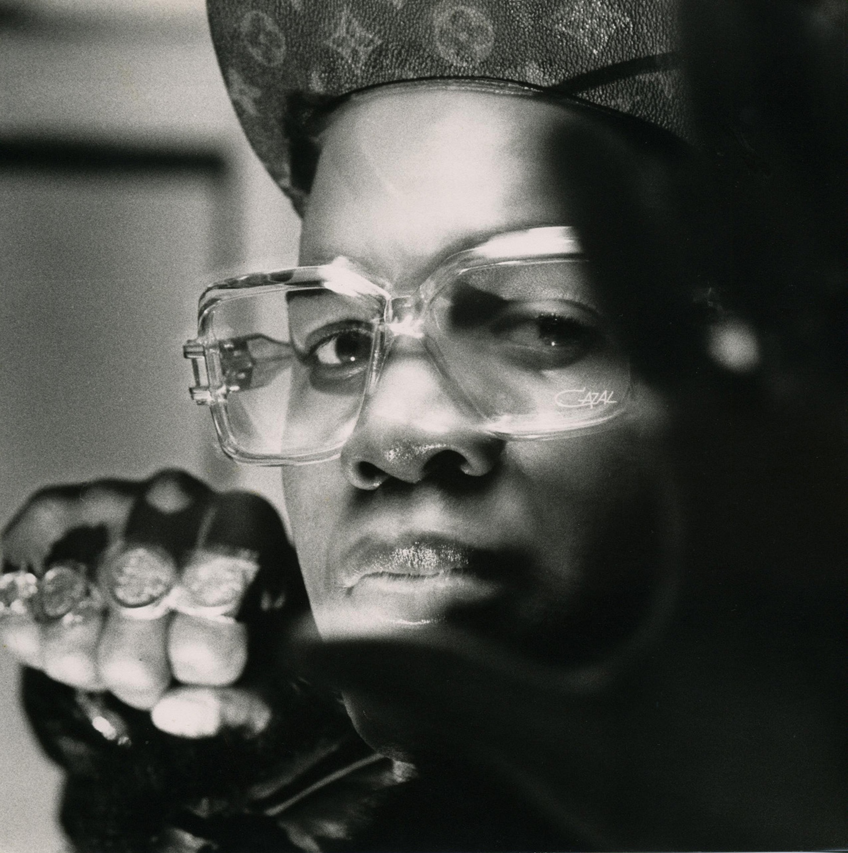 A rapper poses for the camera wearing large clear frame spectacles and clenching his fist.