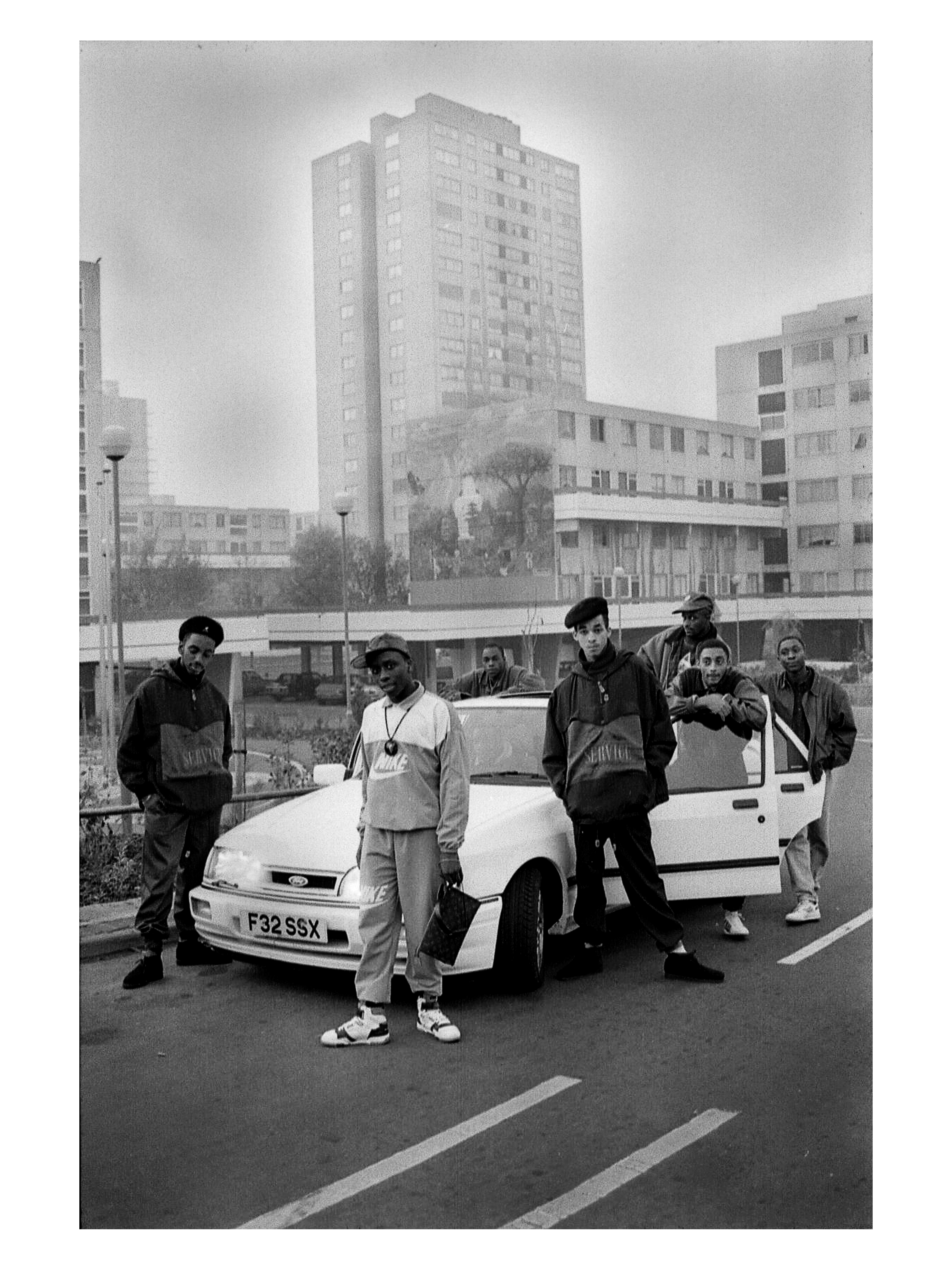 Seven men pose outside of a white car in front of a tower block.