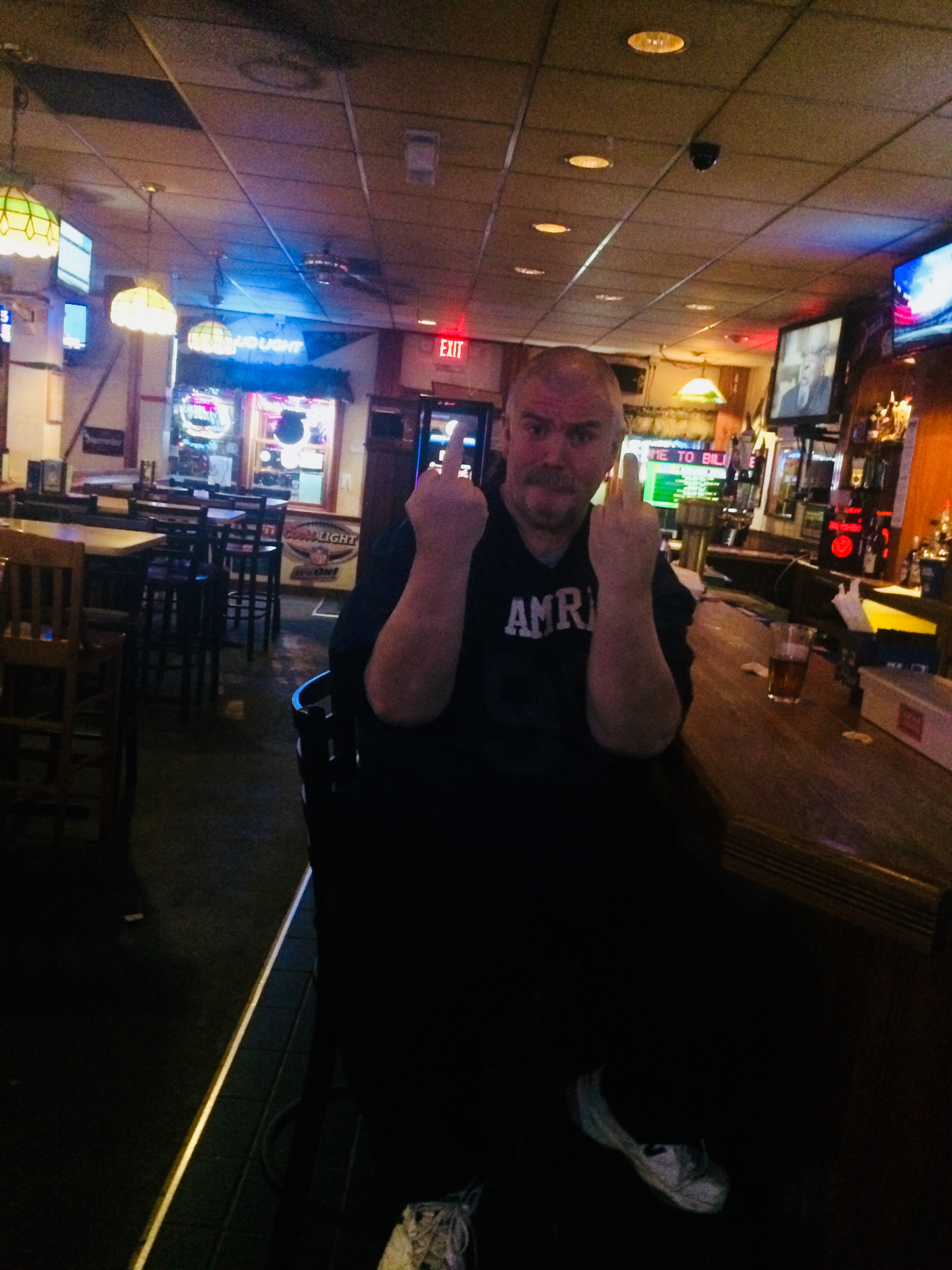 A picture of man in a bar sat on a stool and showing his middle fingers to the camera.