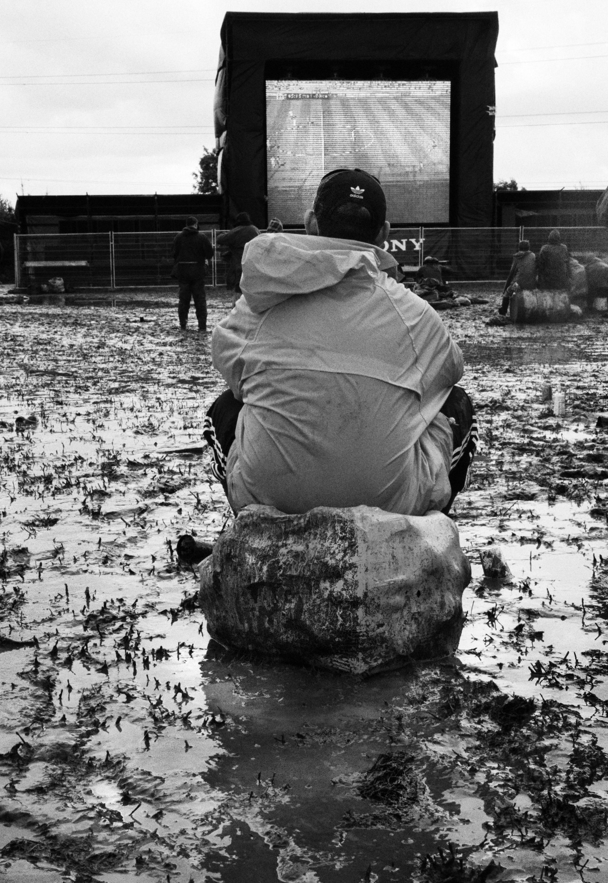 A man sits on a bin bag in a muddy field and watches a big screen.