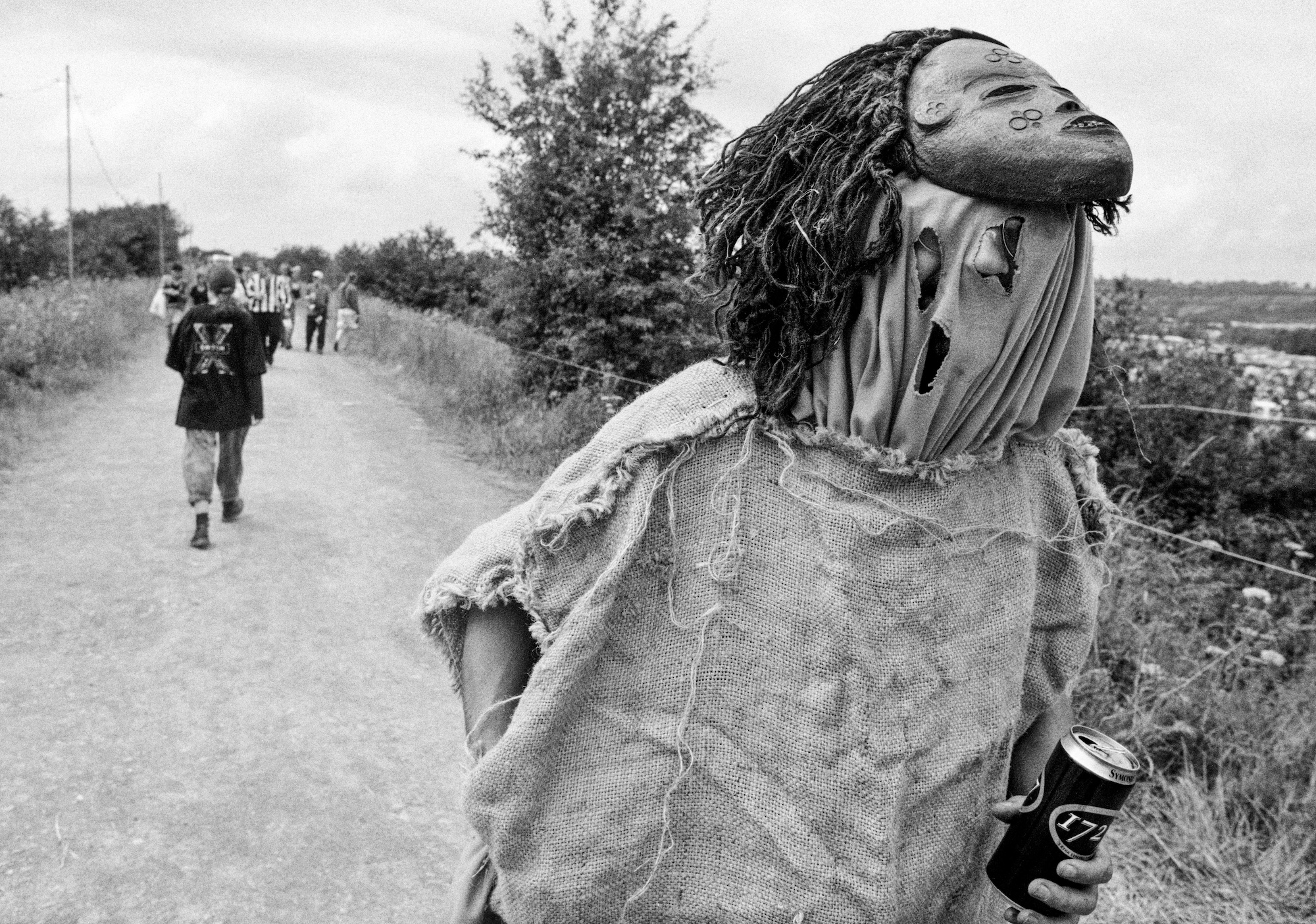 A man in a sack costume and mask walks down a lane carrying a beer.