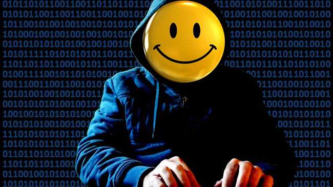 Person in a hoodie with a fake yellow smiley face covering their face as they use a keyboard.