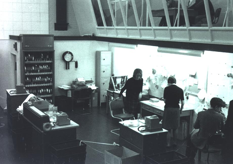 A man and woman in a blue-tinged black-and-white photograph stand in front of a bulletin board lit by fluorescent strip lights in the old Cambridge Mathematical Laboratory, beneath a row of viewing gallery windows. The woman is speaking as she rises from her chair.