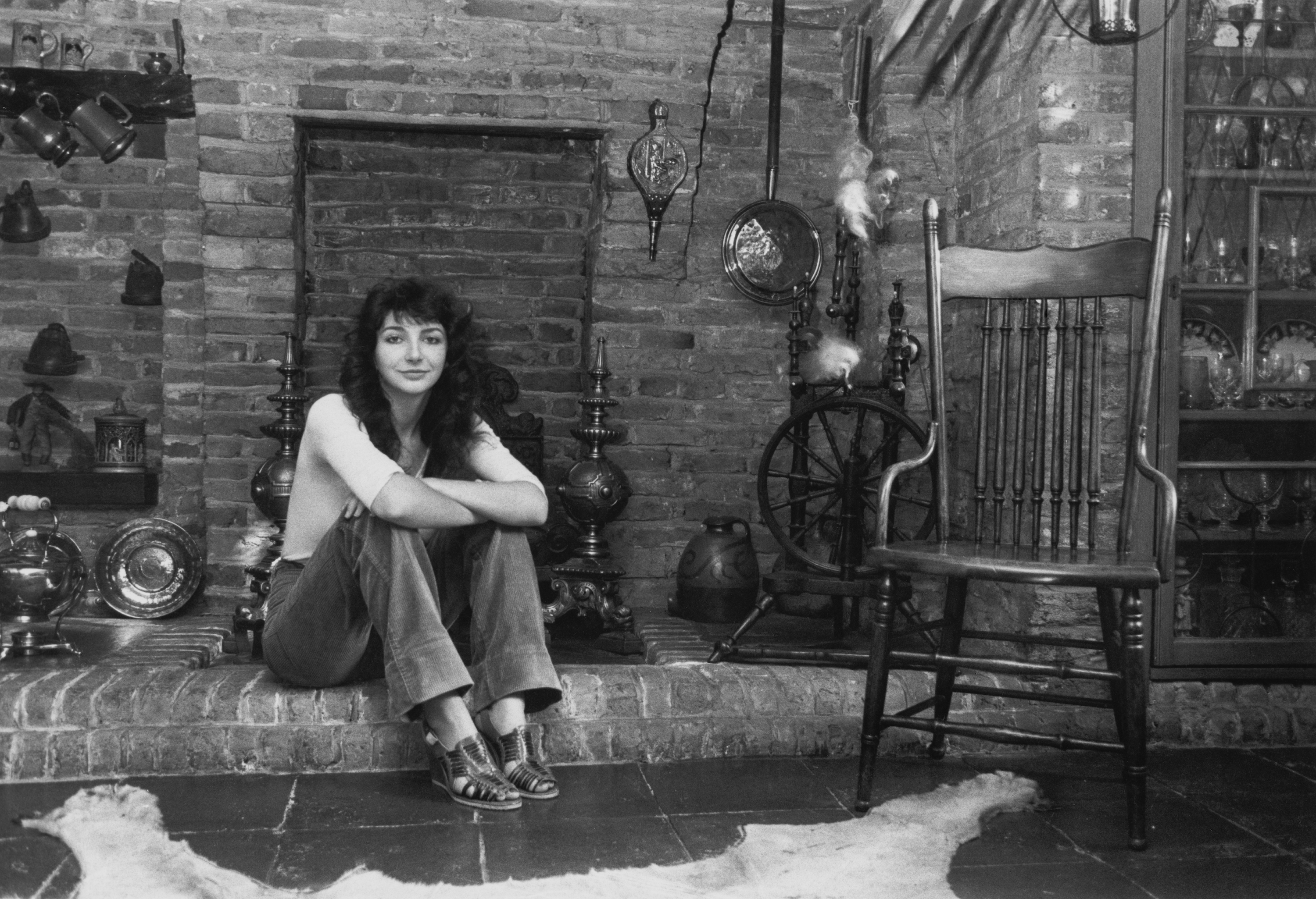 Kate Bush sitting on a fireplace at her family home in 1978, surrounded by pots, jugs, a spinning wheel, wooden chair and bear rug while wearing jeans, a white fitted crop top and wedged sandals.