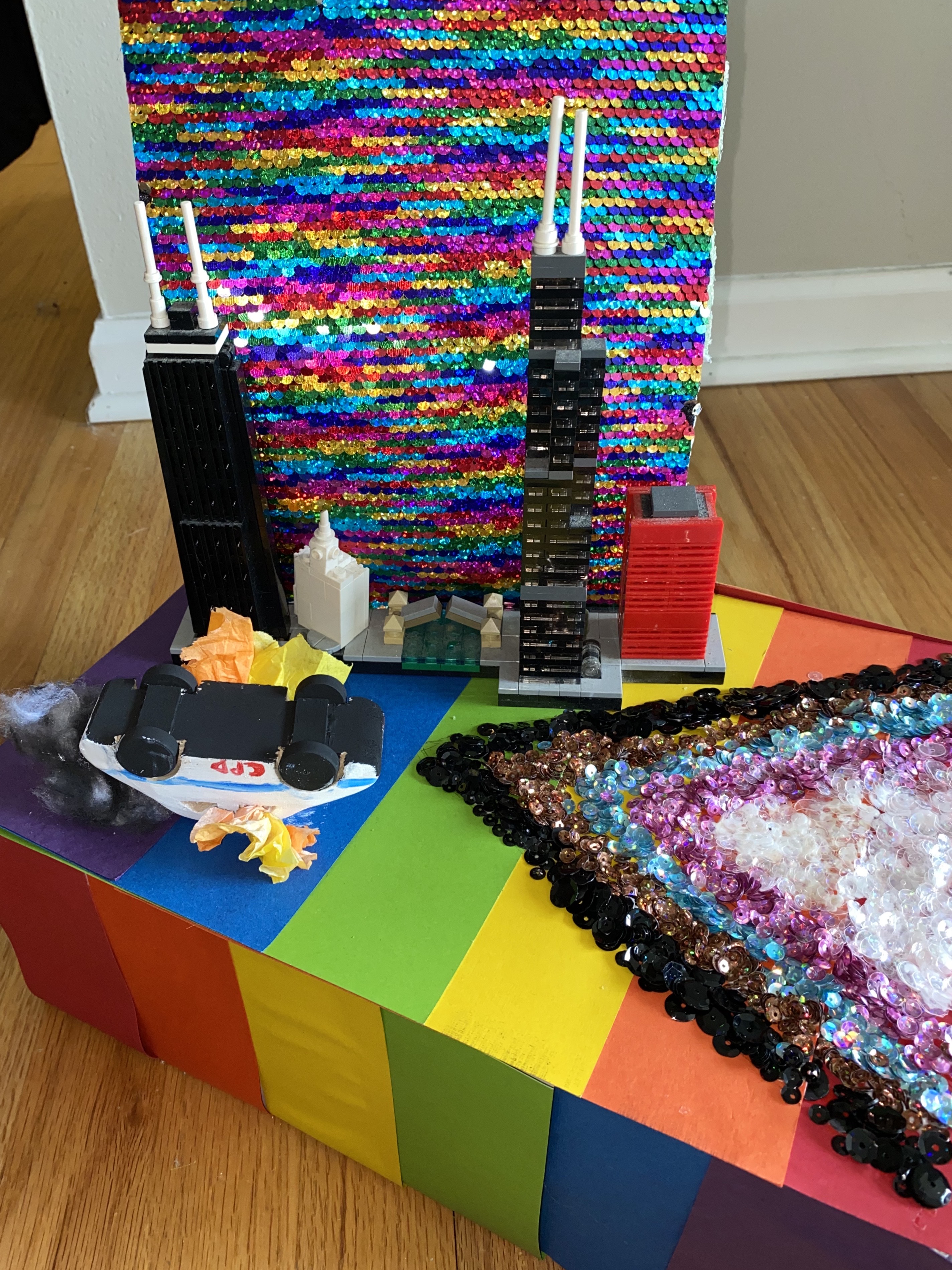 miniature pride float featuring the chicago skyline, a trans flag with black and brown lines added, and a burning cop car